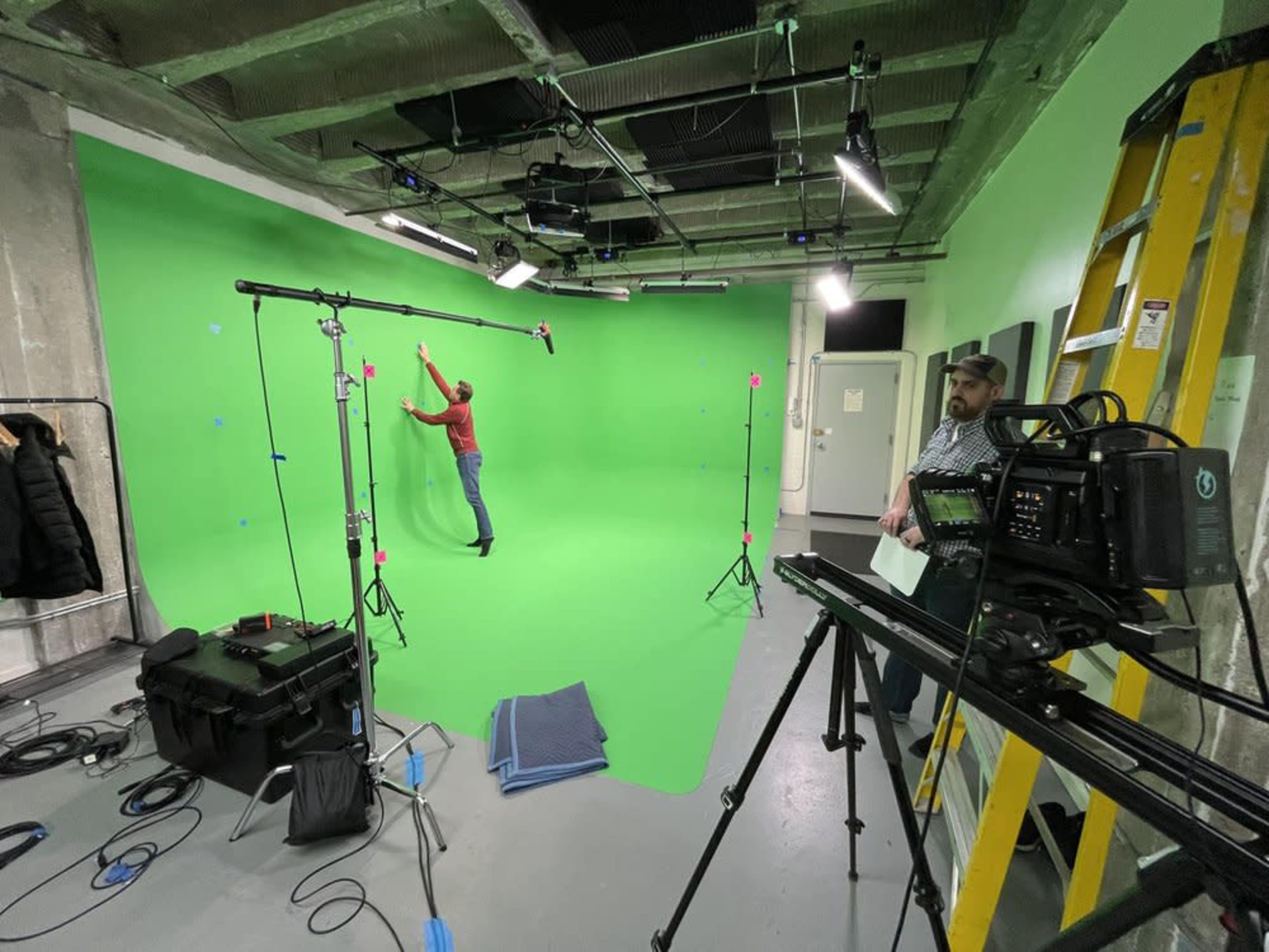 A man performs in a green screen studio while another man operates a camera on a tripod.
