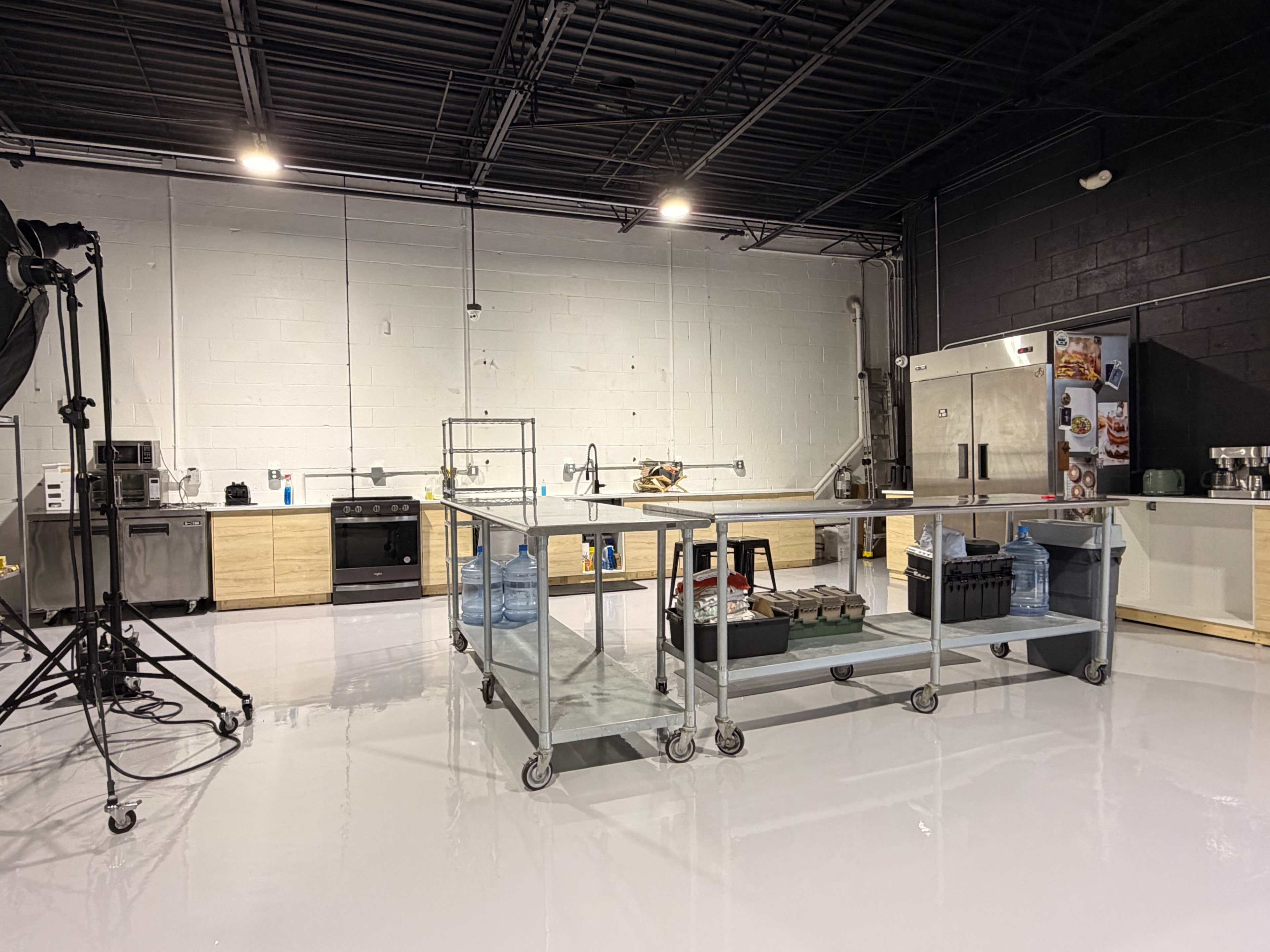 The image shows a modern kitchen studio with stainless steel appliances, a large central workspace on wheels, and a clean, glossy floor.