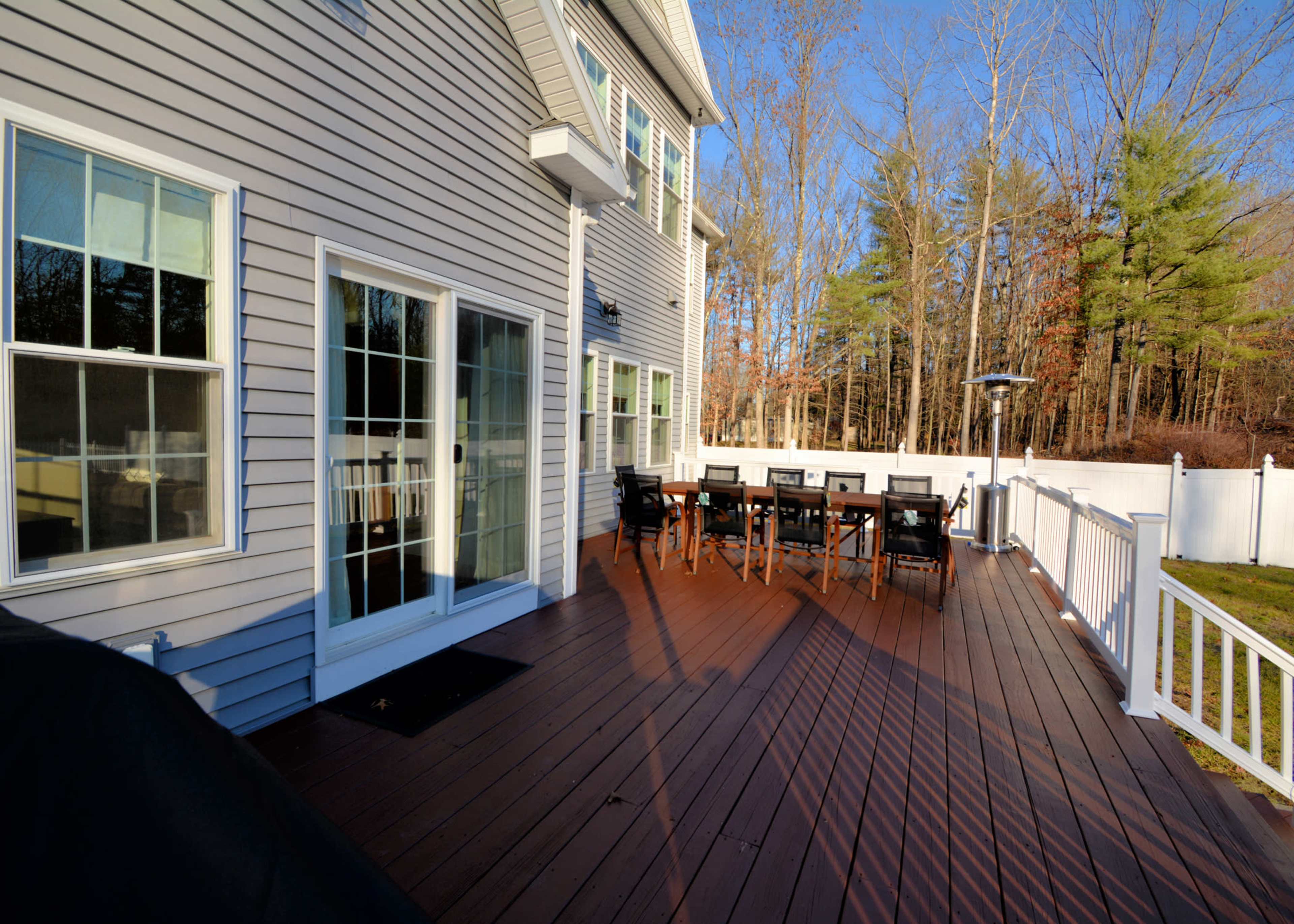 A wooden deck adjacent to a gray house, with a dining set and a backdrop of trees.