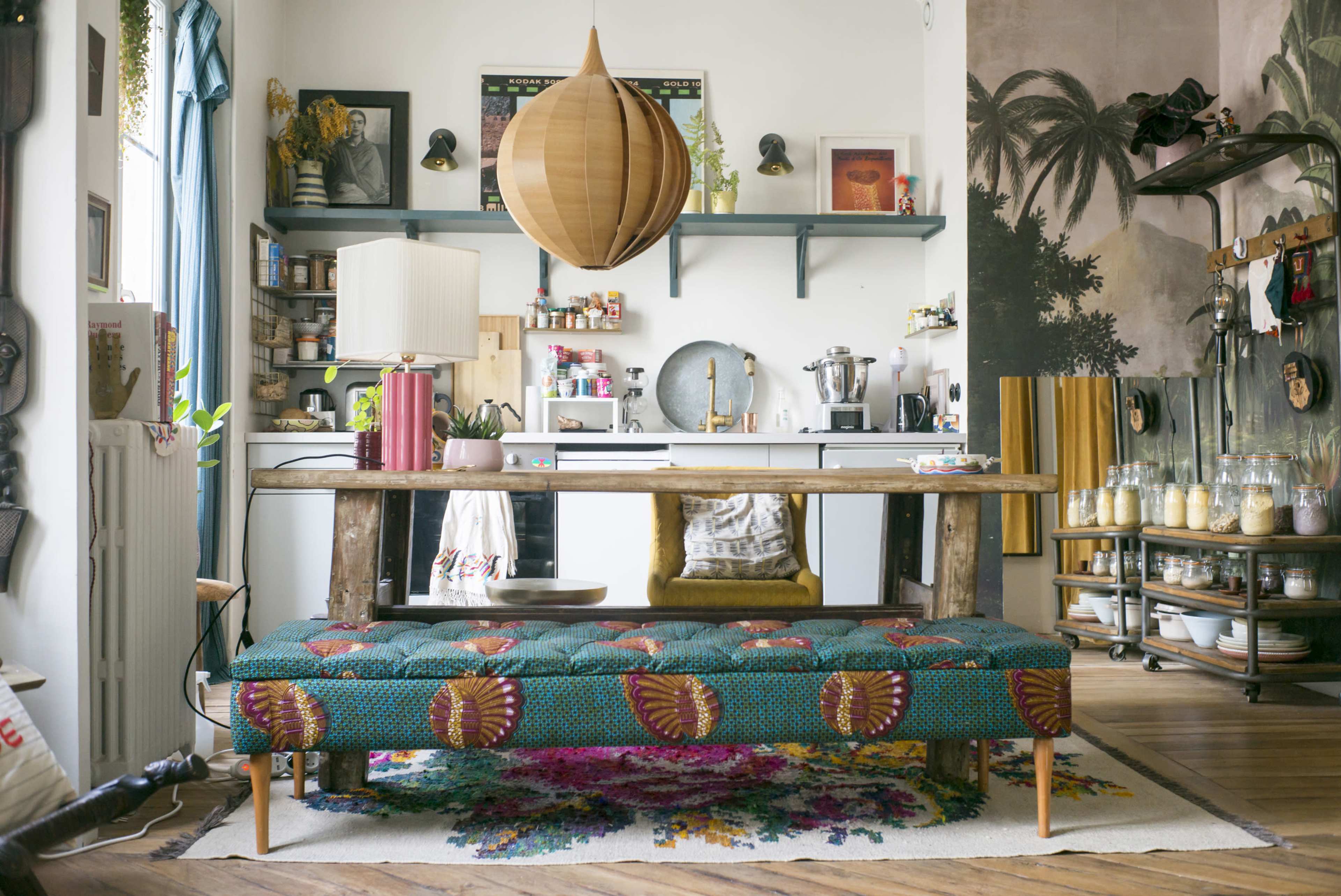 A kitchen and dining area features a large wooden table, a decorative bench with vibrant fabric, and various kitchen items displayed on shelves.