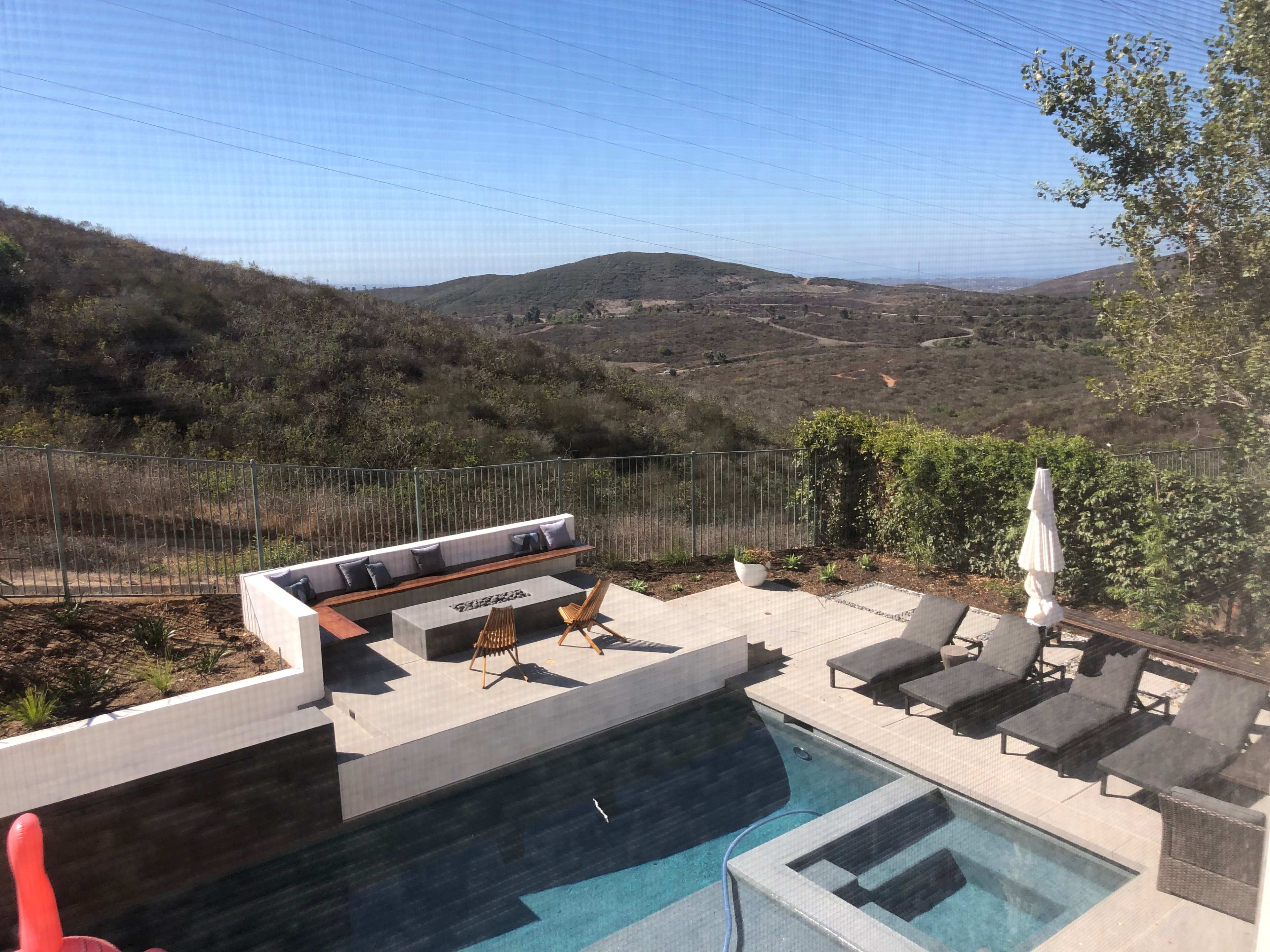 The image shows a patio with a pool, lounges, and a fire pit, overlooking a mountainous landscape on a clear day.