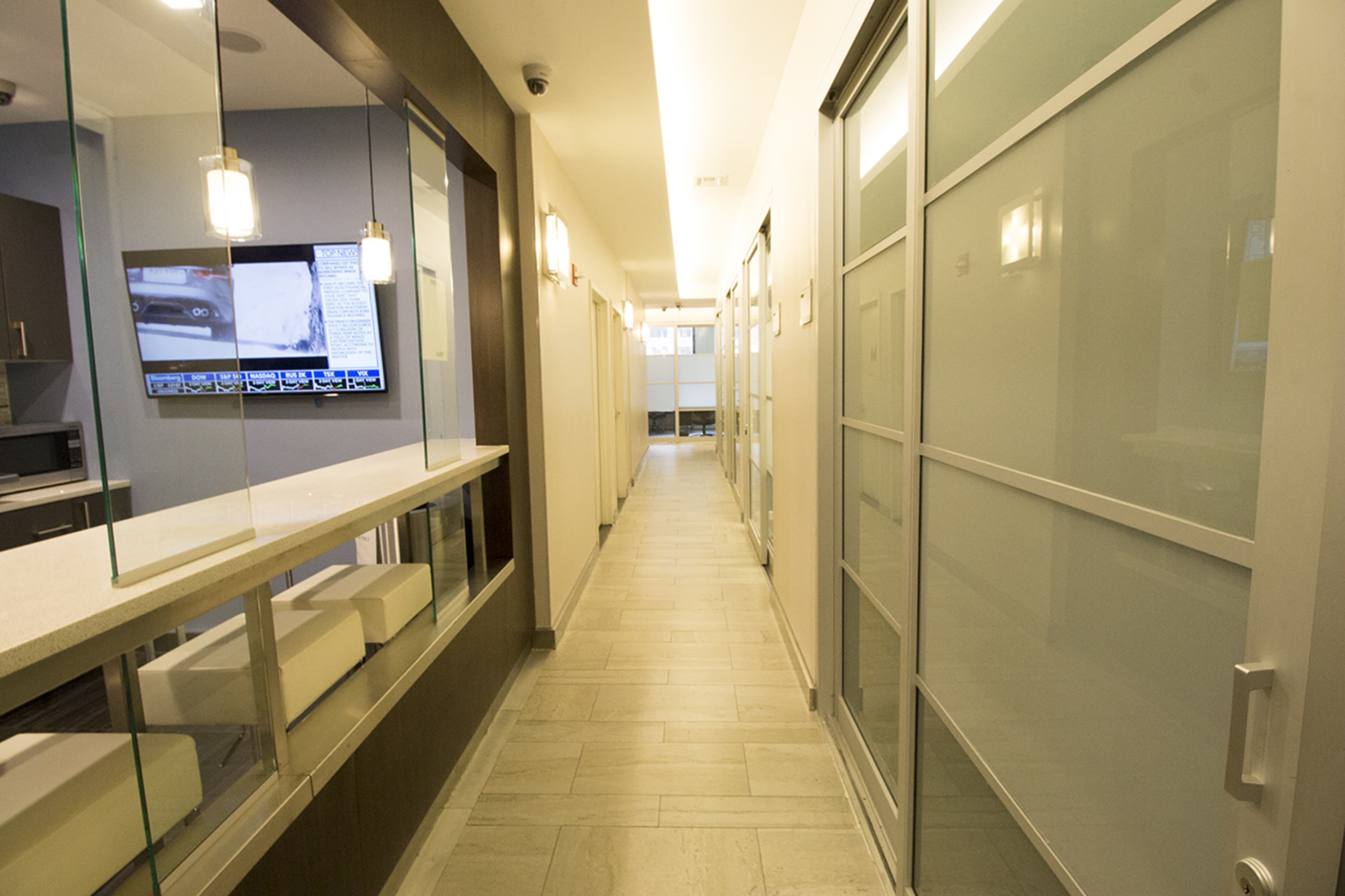A modern hallway features glass-enclosed offices on either side and a television mounted on the wall at the end.