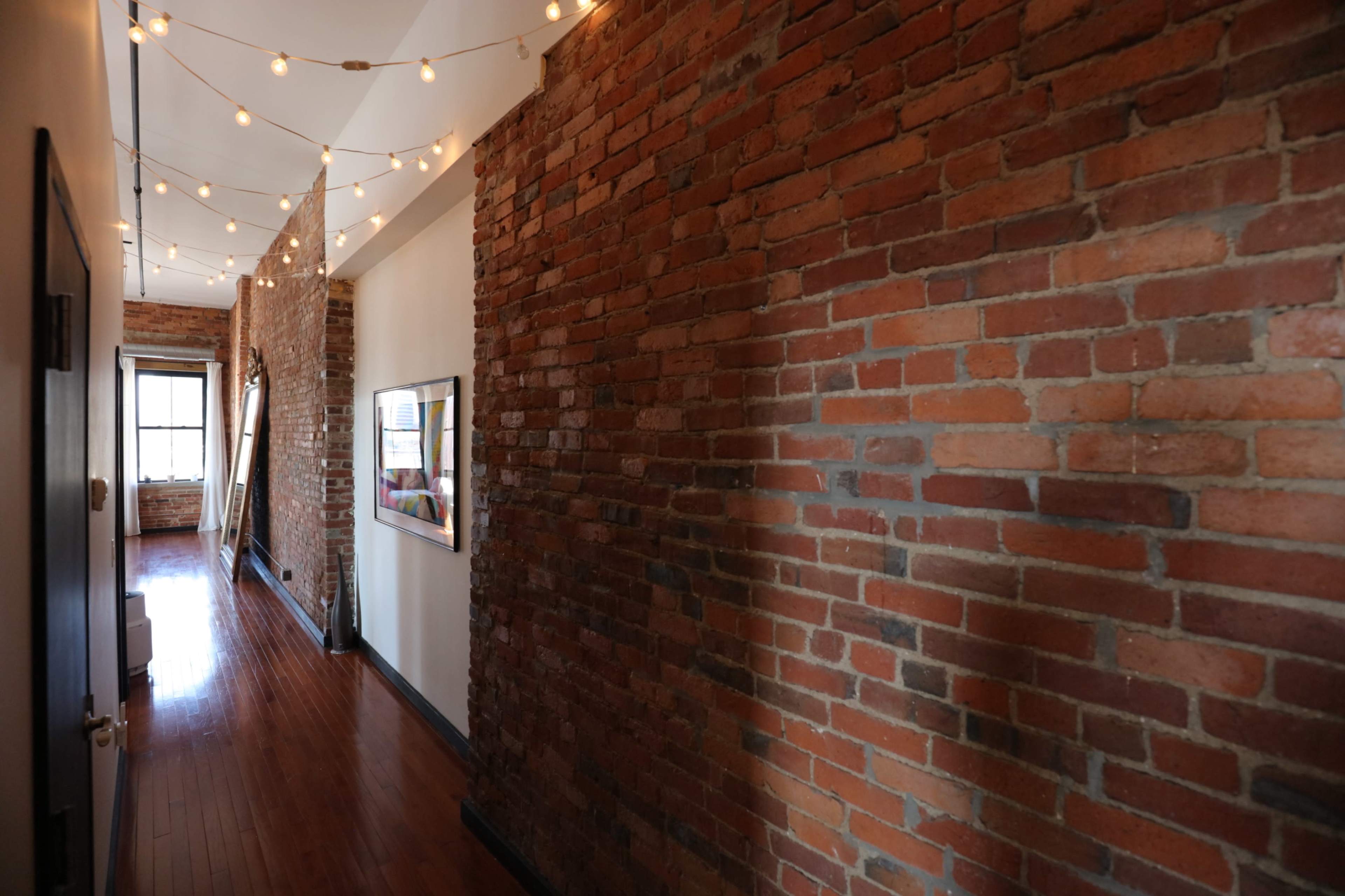 The image shows a narrow hallway with exposed brick walls and string lights overhead, featuring wooden flooring and a framed picture on one side.