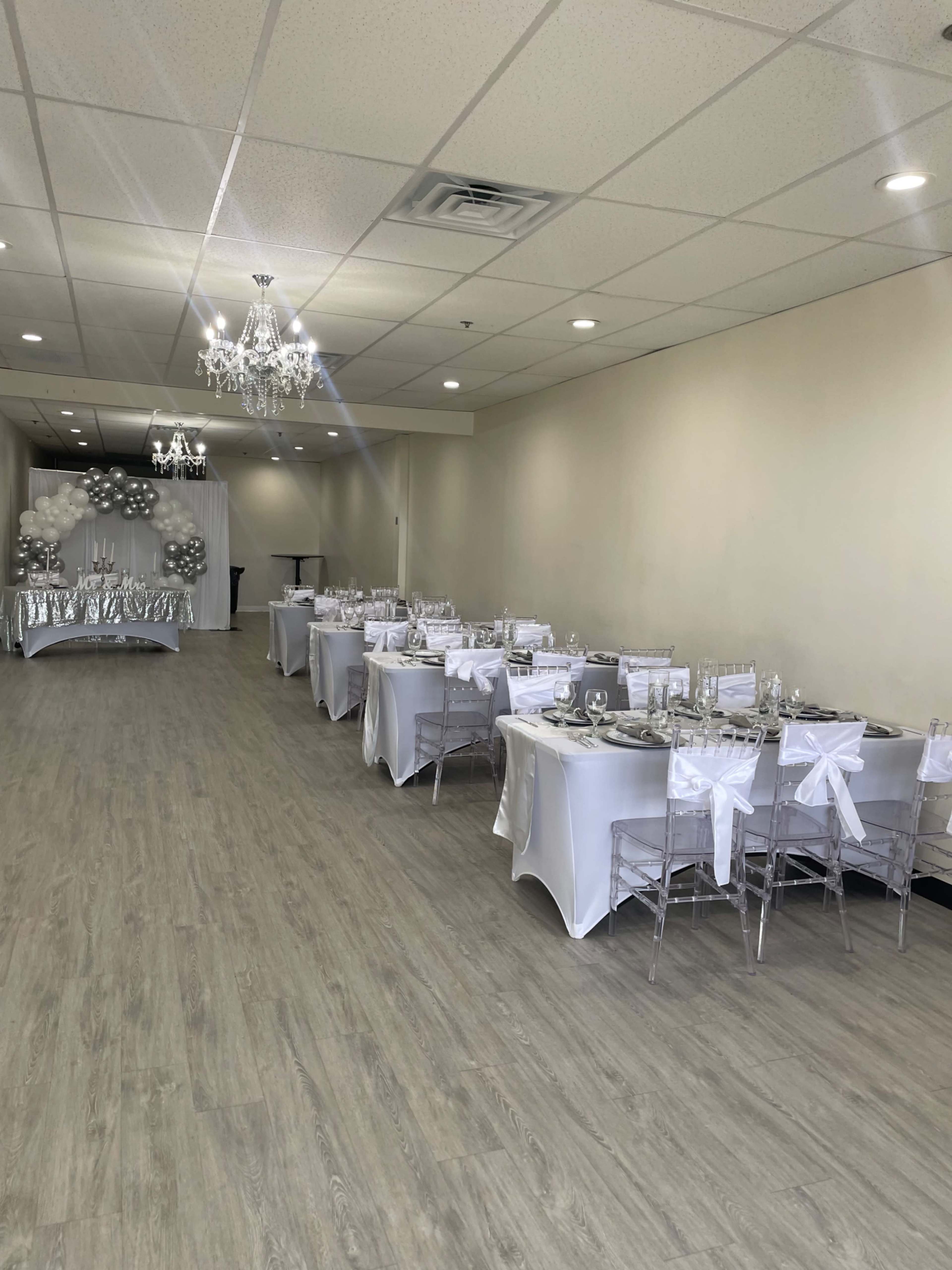A decorated event space features white tables with elegant place settings, clear chairs, and chandeliers, creating a formal atmosphere for gathering.