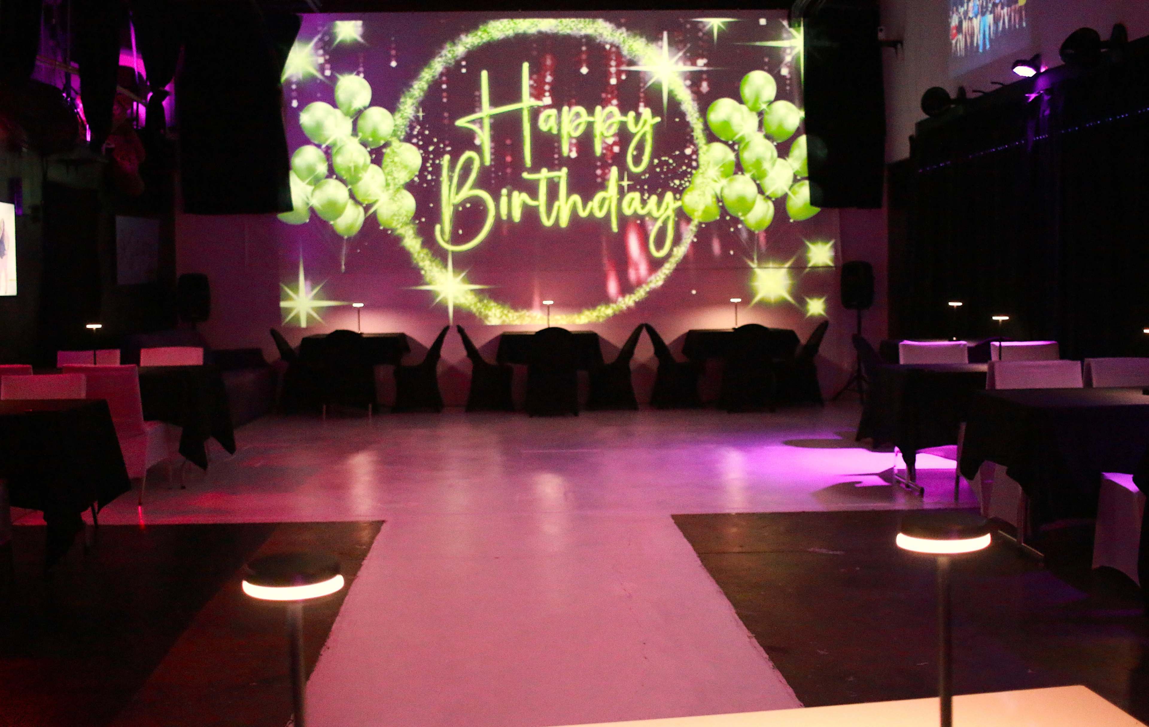 The image shows a decorated event space with black tables, purple lighting, and a large screen displaying "Happy Birthday" with green balloons.