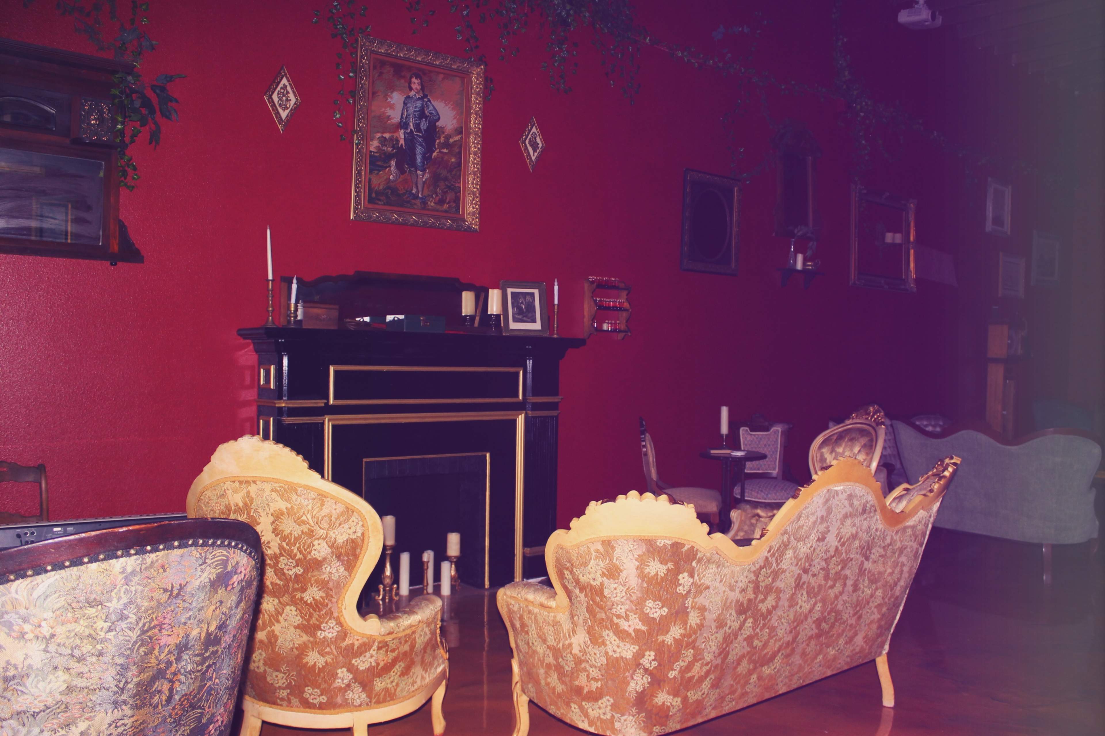 The interior features a red-walled room with antique furniture arranged around a black fireplace, adorned with decorative art and greenery.