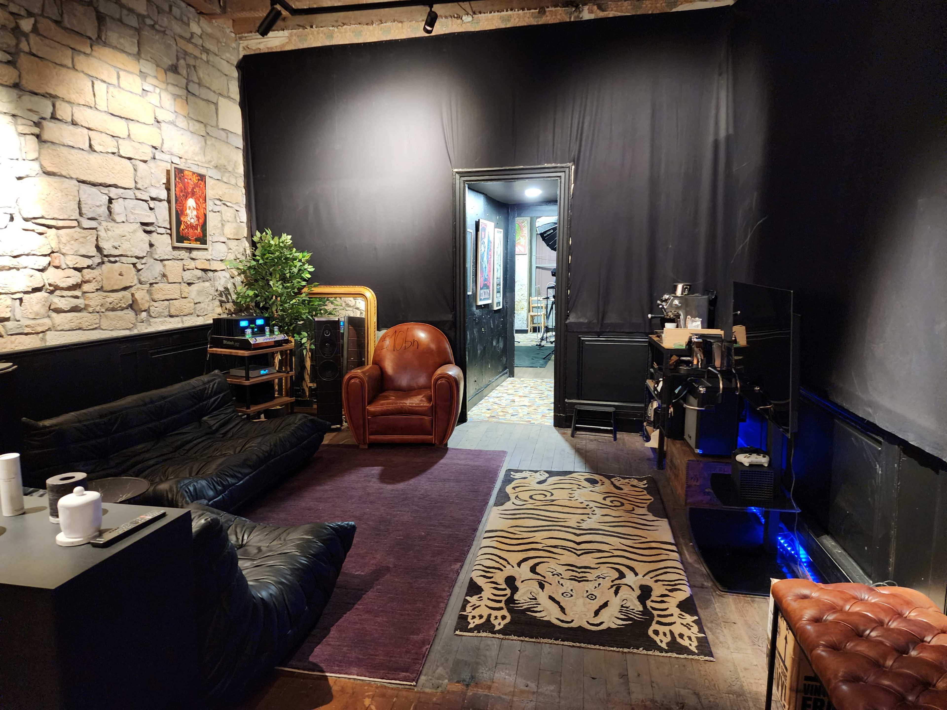 The image shows a dimly-lit room featuring a black wall, a large purple rug, a brown leather armchair, and two black bean bag chairs, along with a black TV stand and a doorway leading to another space.