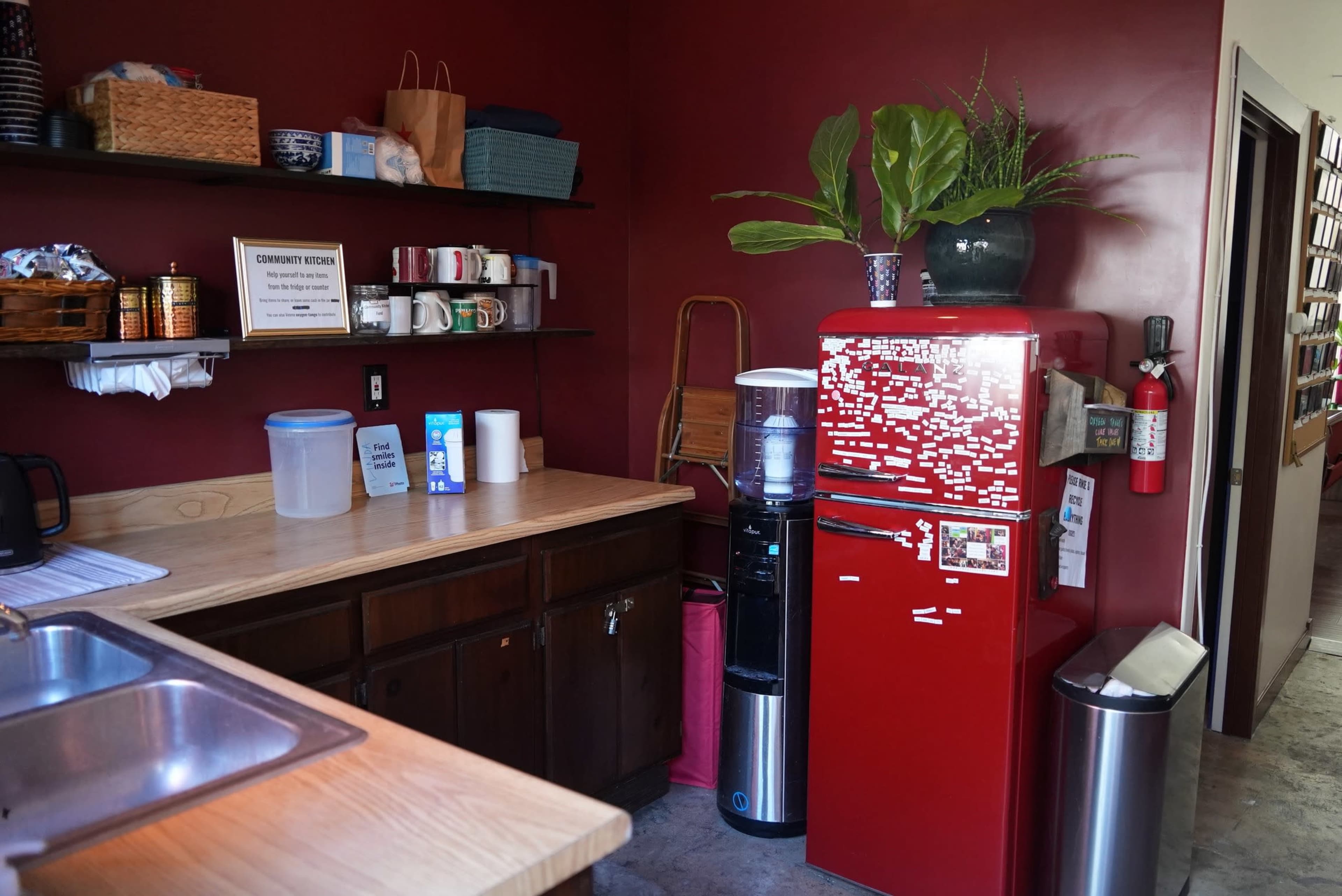 A kitchen with a red refrigerator, wooden shelves holding various items, a sink, and a water cooler against a dark red wall.