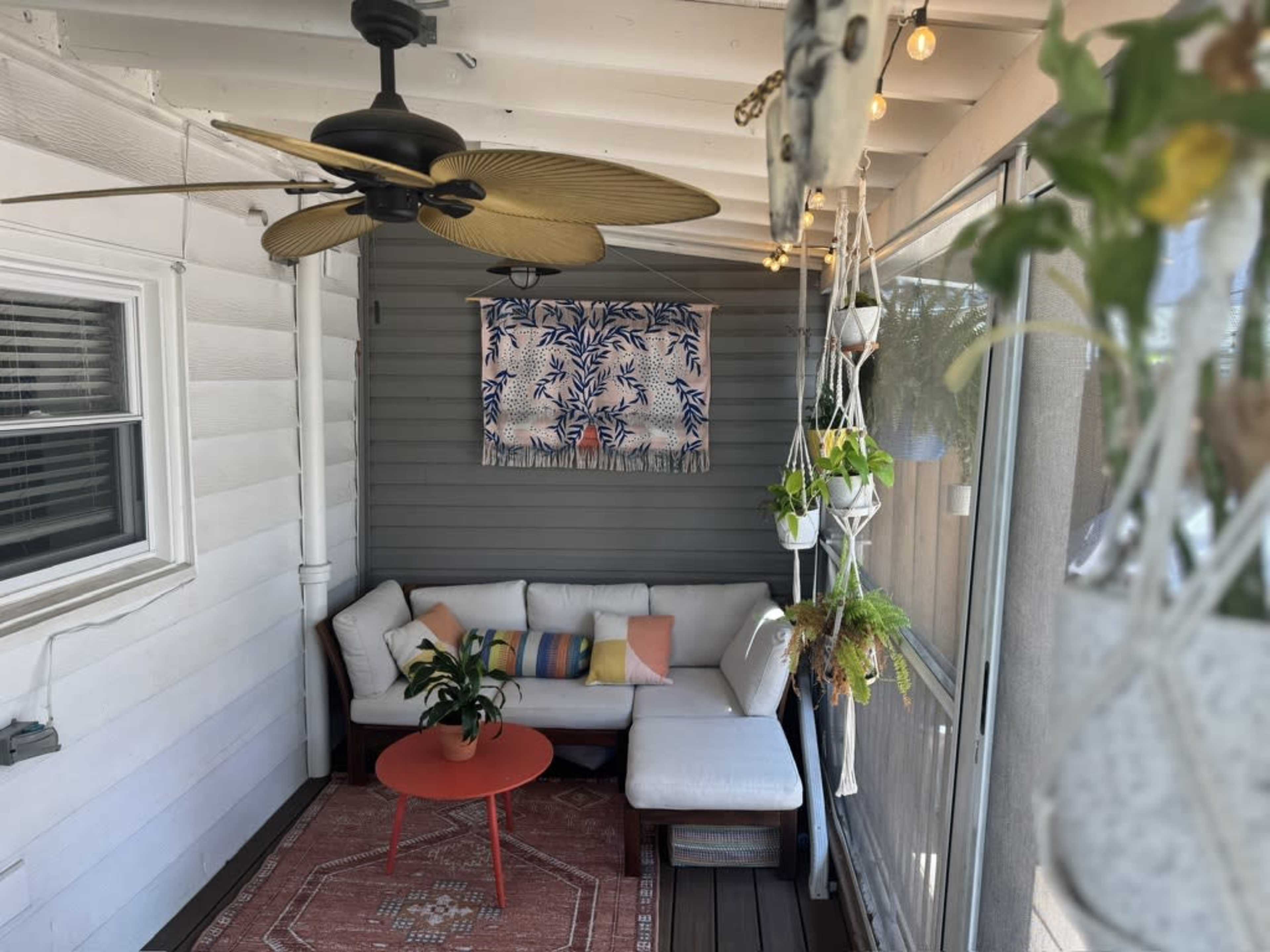 The image shows a cozy porch area featuring a sectional sofa with colorful cushions, a red coffee table, and plants hanging from the ceiling.