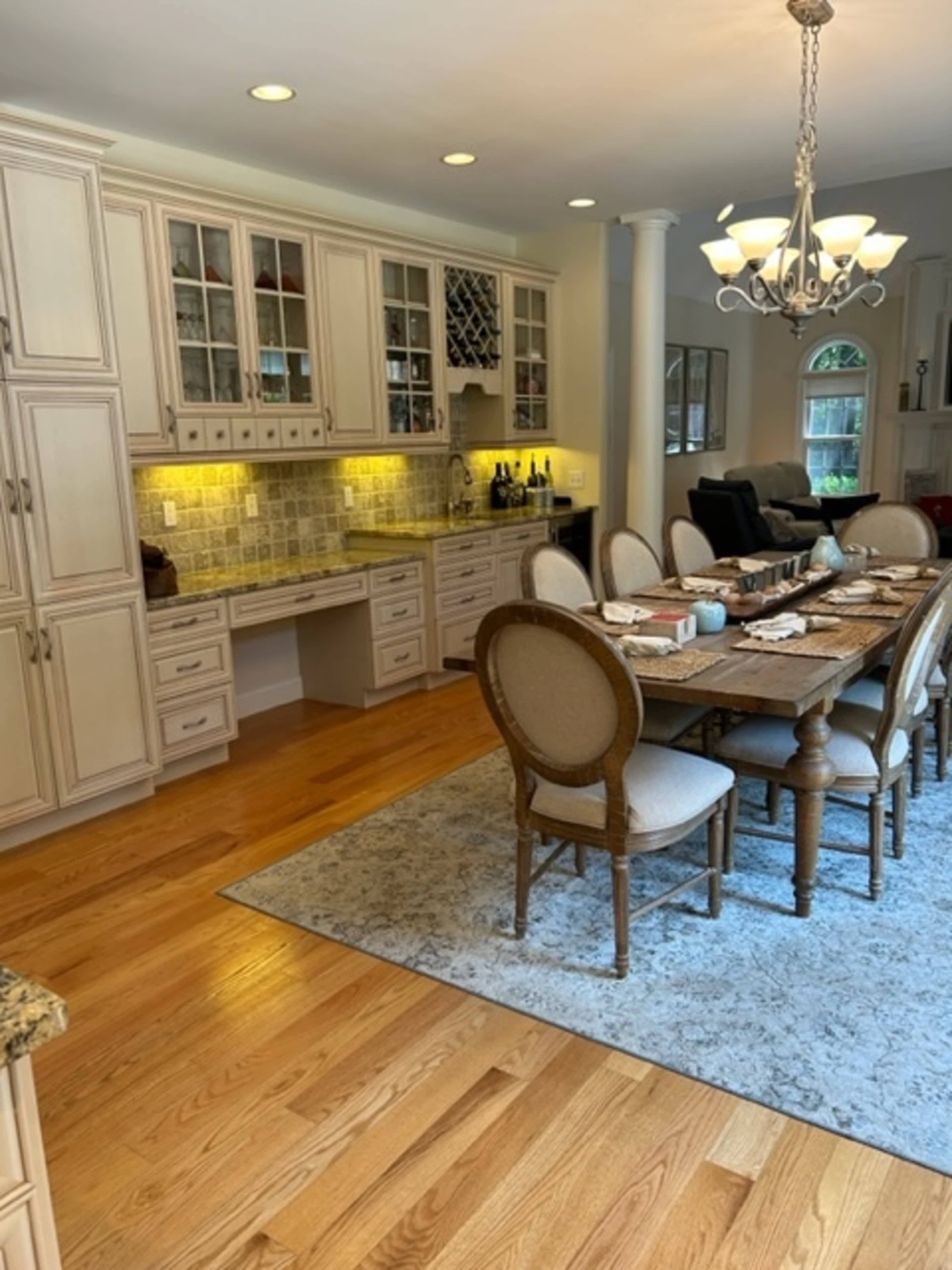 A spacious kitchen features a long dining table surrounded by upholstered chairs, with cabinets above and a countertop illuminated by under-cabinet lighting.