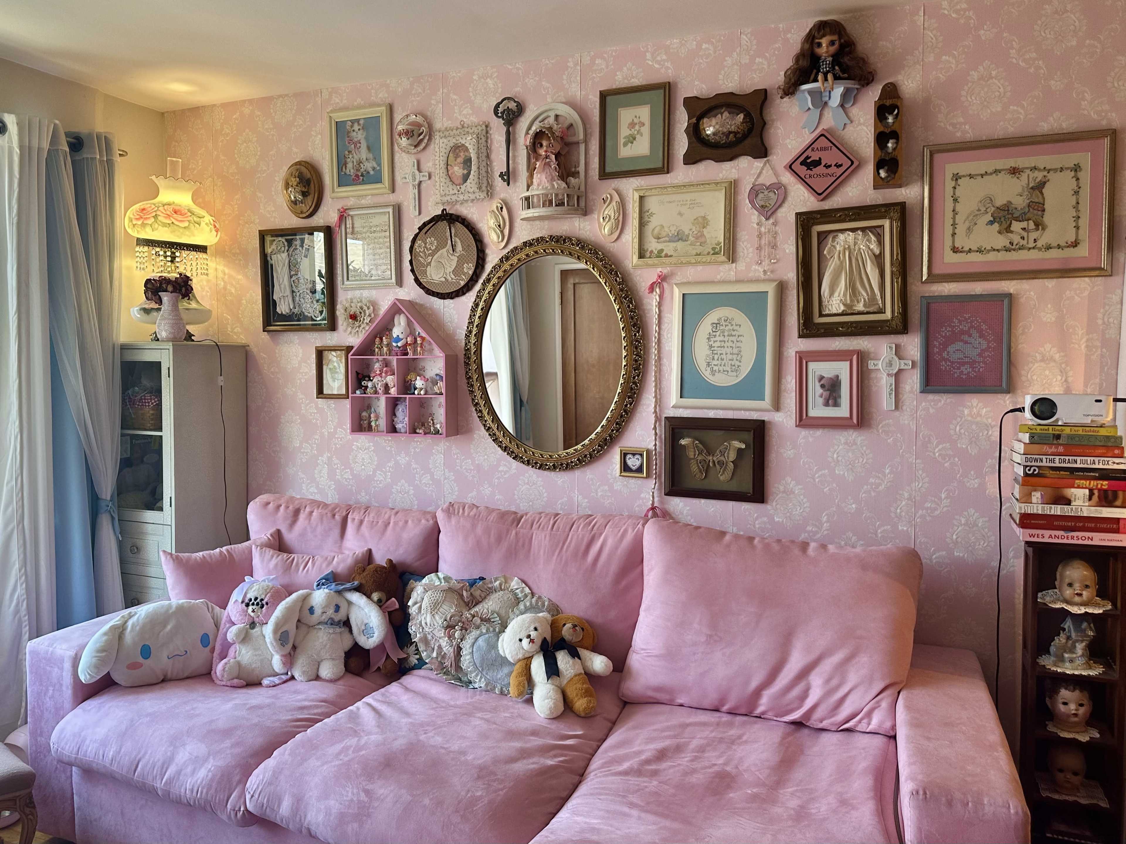 The scene features a pink sofa adorned with stuffed animals, against a wall decorated with an arrangement of framed pictures and mirrors.