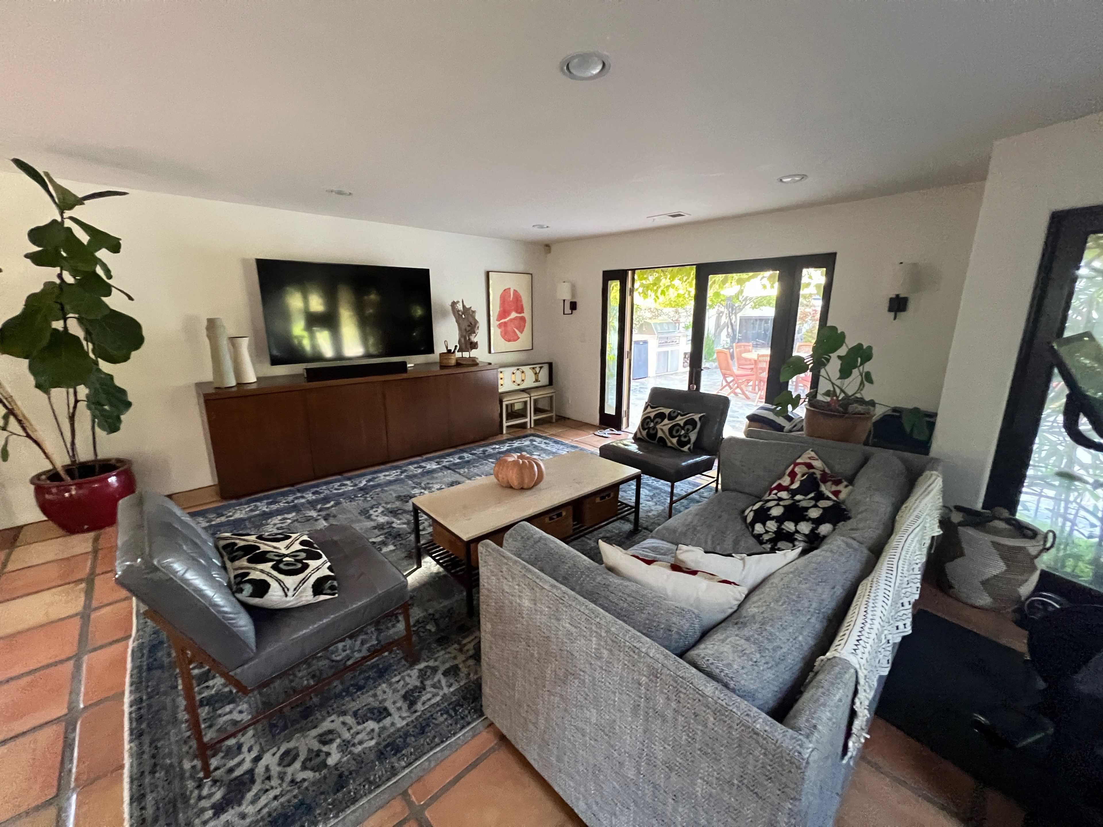 The image shows a modern living room with a television, a coffee table, and seating arranged around a patterned rug, featuring large windows that let in natural light.