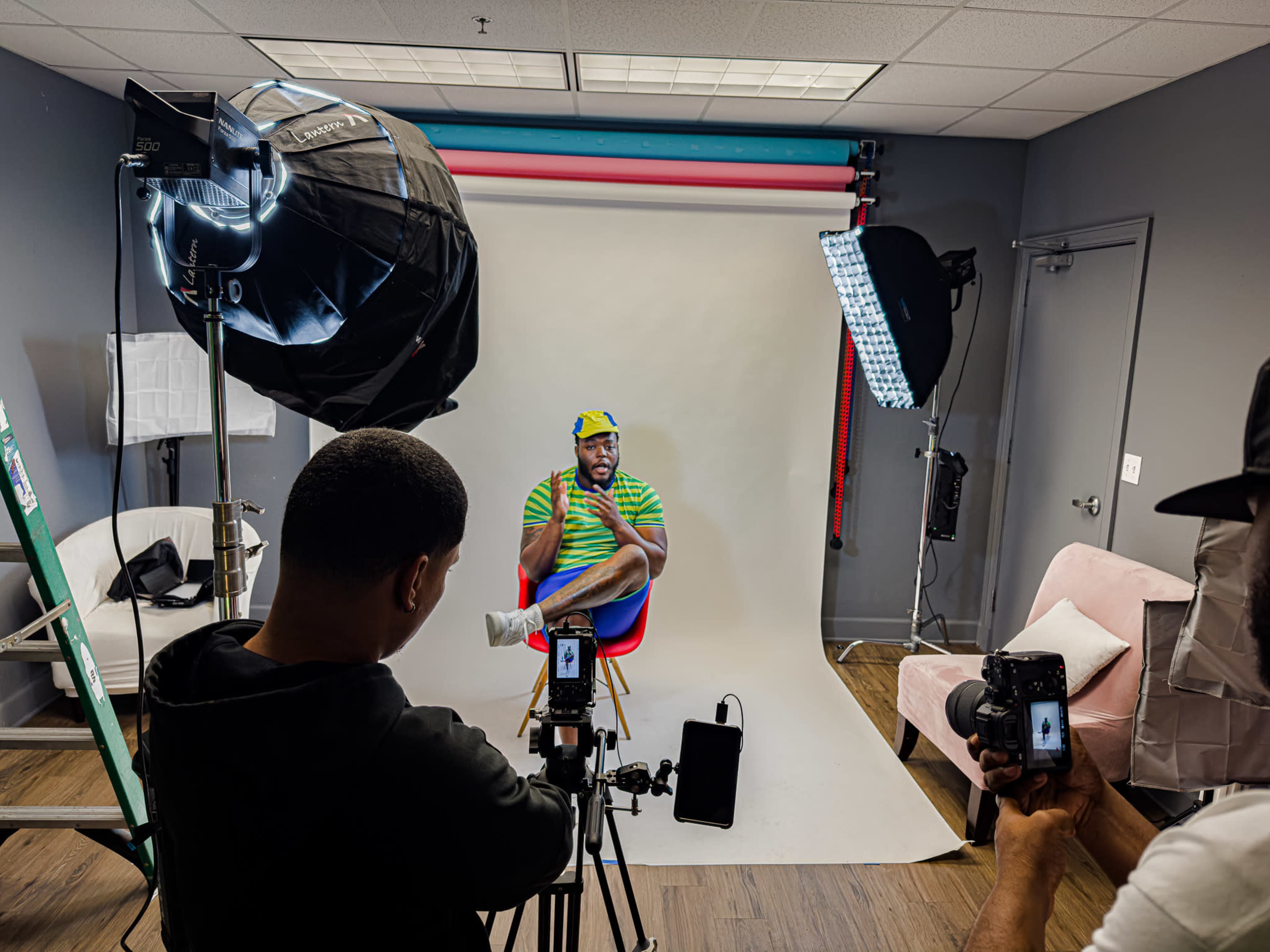 A man sits on a stool in front of a backdrop while being photographed by two individuals holding cameras in a well-lit studio.