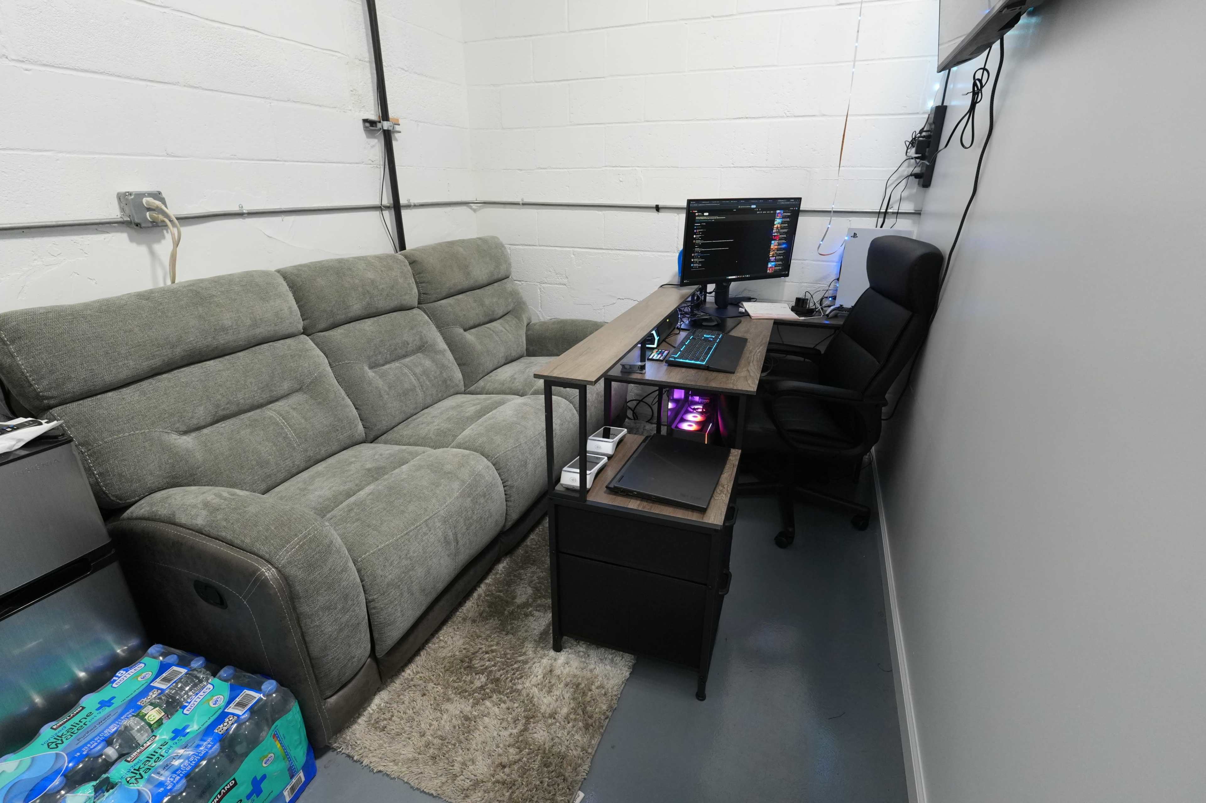 The image shows a small office space featuring a gray couch, a desk with a computer monitor, and a black office chair against a white brick wall.