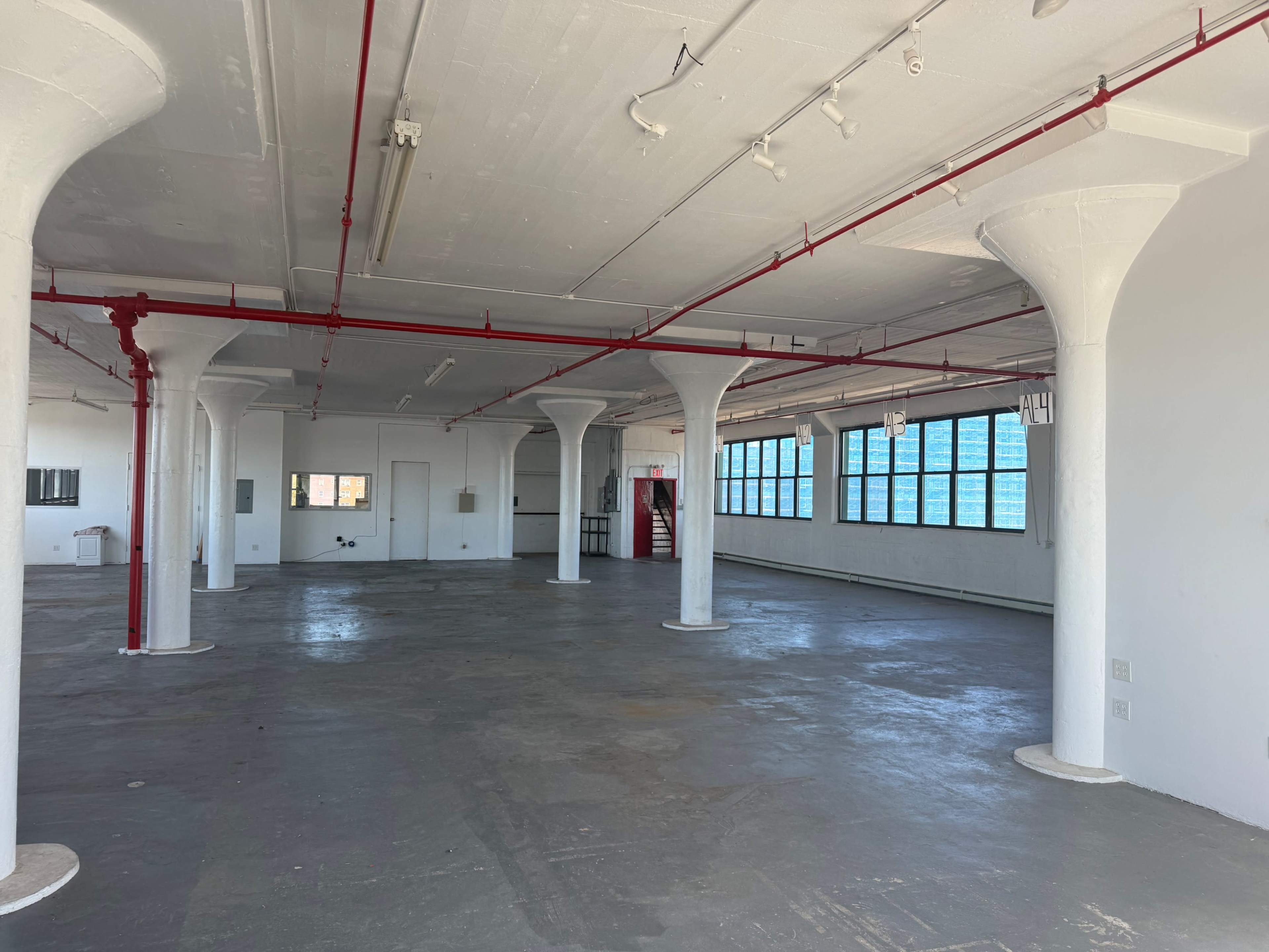 The image shows a large, empty industrial space with exposed columns, red piping, and large windows.