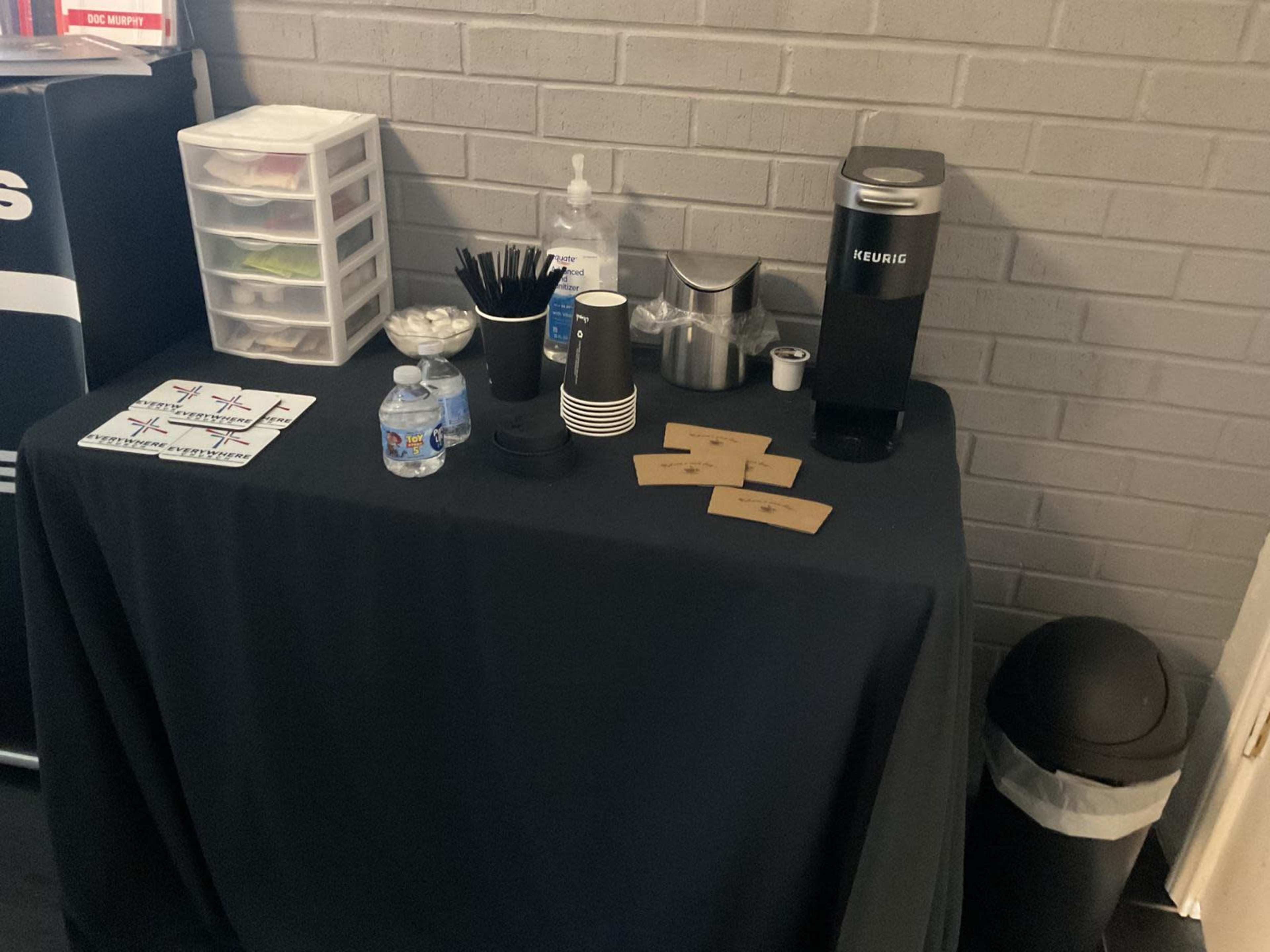 A table with a Keurig coffee maker, cups, water bottles, and various supplies is set up against a gray brick wall.
