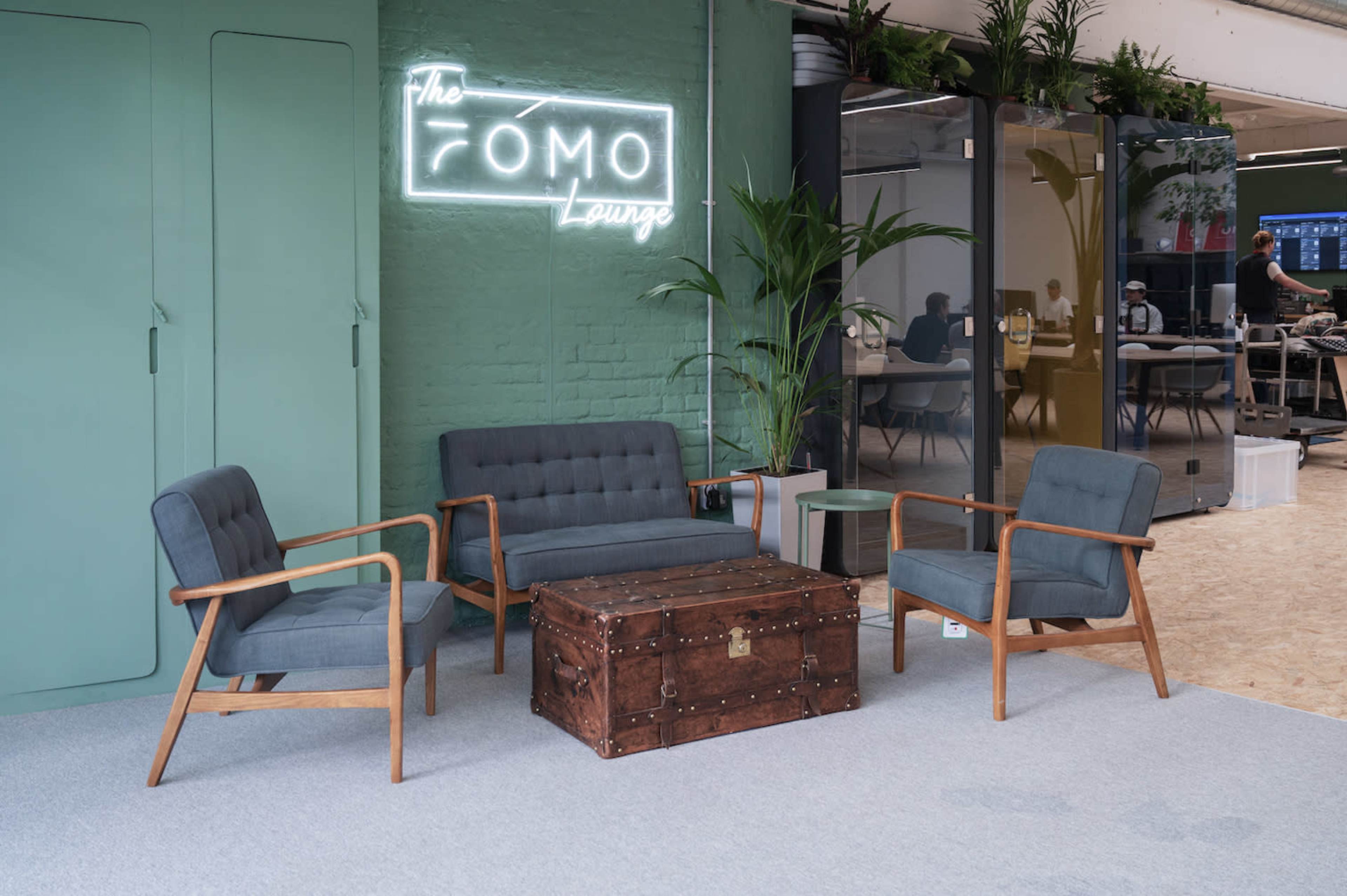 The image shows a cozy lounge area with a wooden coffee table, three upholstered chairs, and a neon sign that reads "the FOMO Lounge" against a green wall.