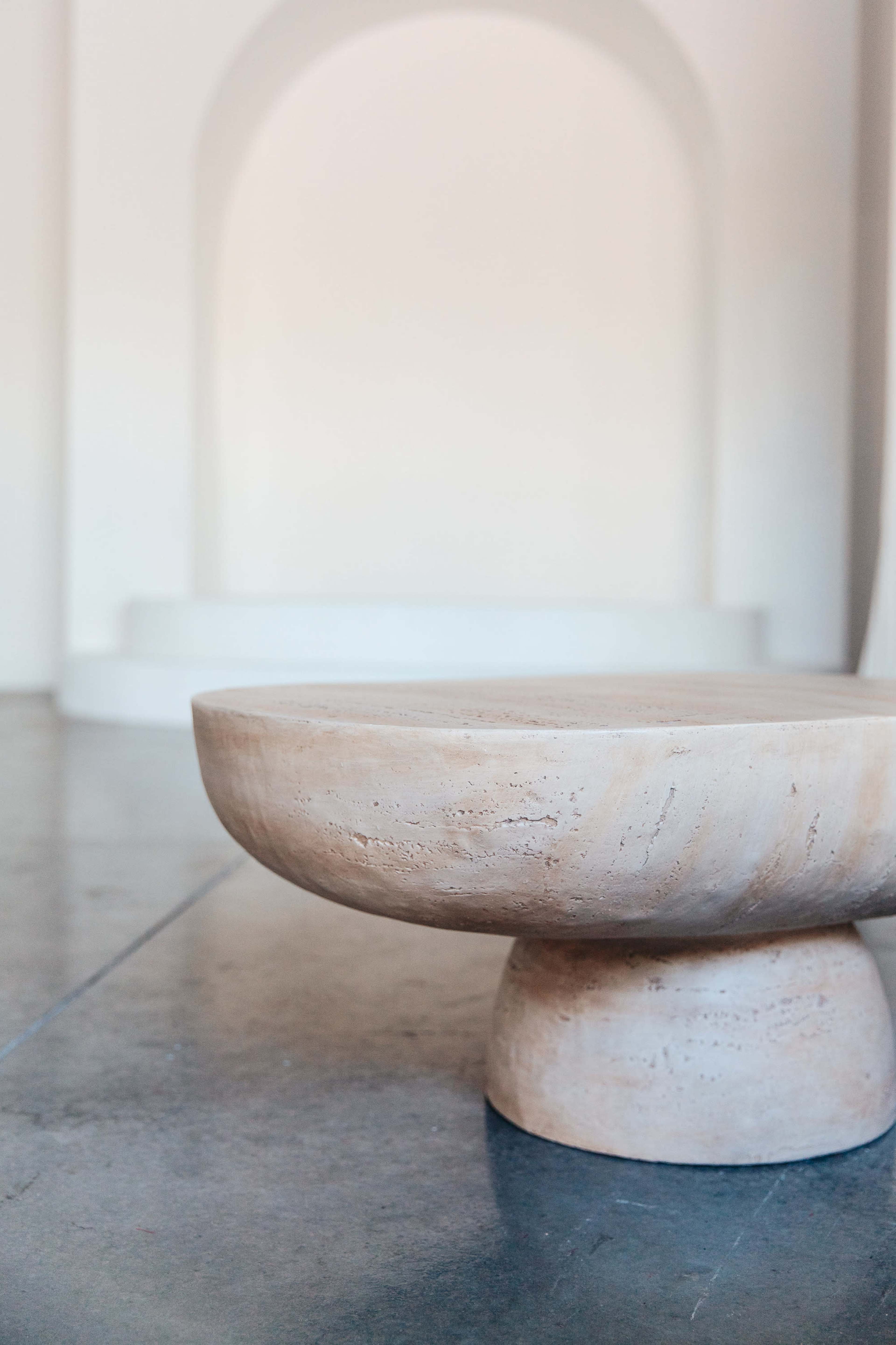 A round, textured stone table is positioned in the foreground, with a minimalist white space and archway visible in the background.