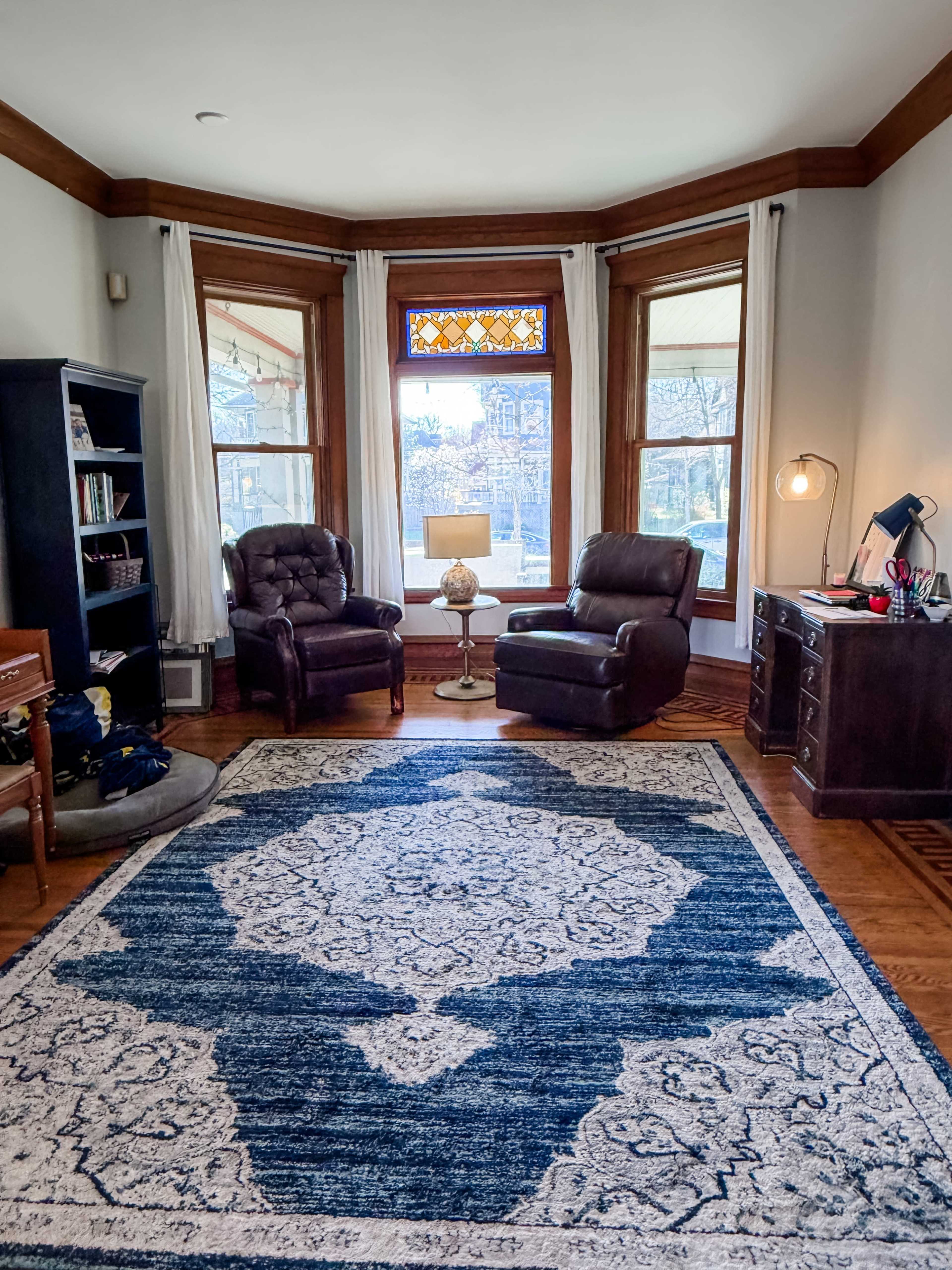 The image shows a living room with two recliner chairs facing a window, a round lamp on a small table between them, and a desk against one wall, all situated on a large blue patterned rug.