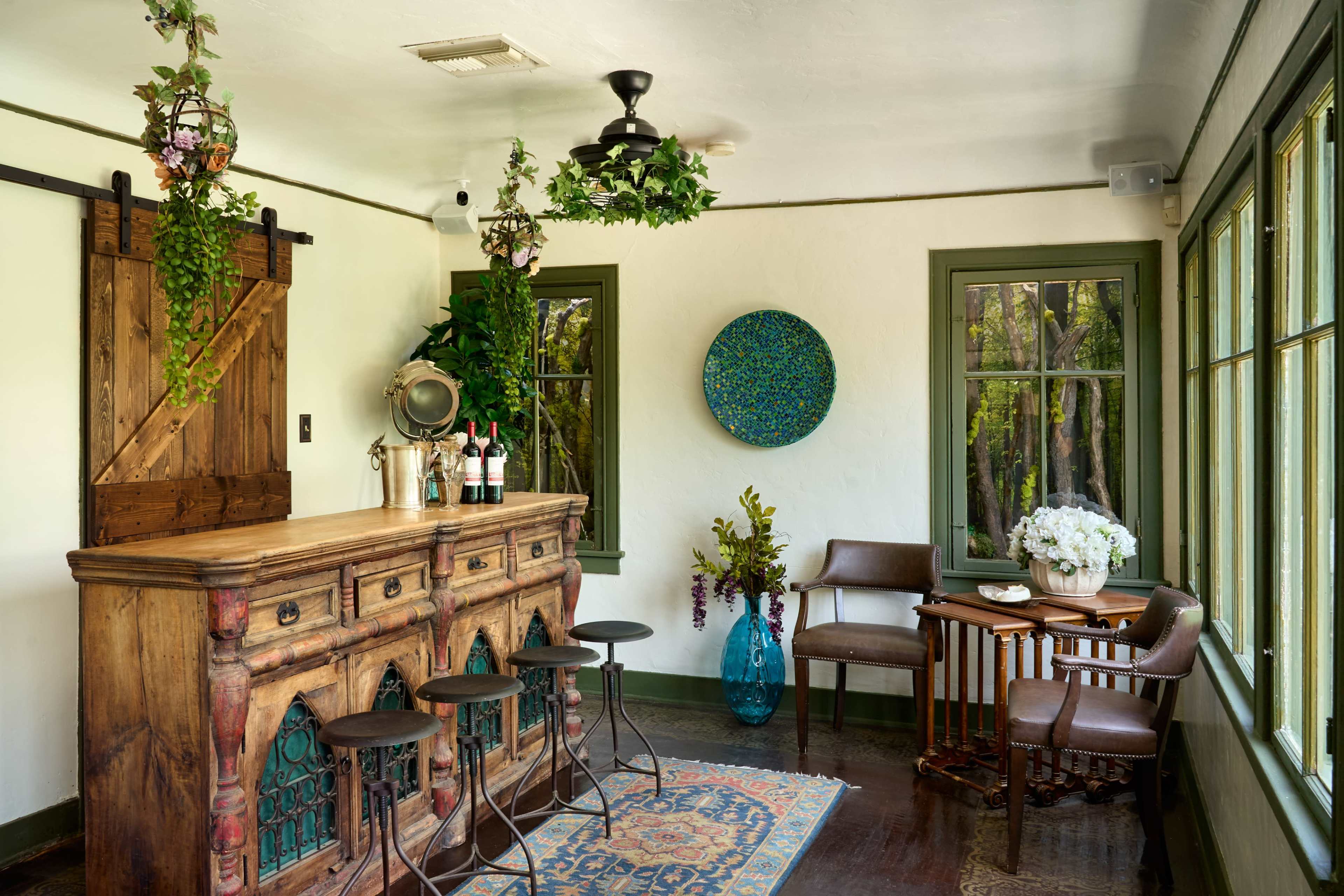 A cozy room featuring a wooden bar with stools, large windows, and various decorative elements including plants and artwork.