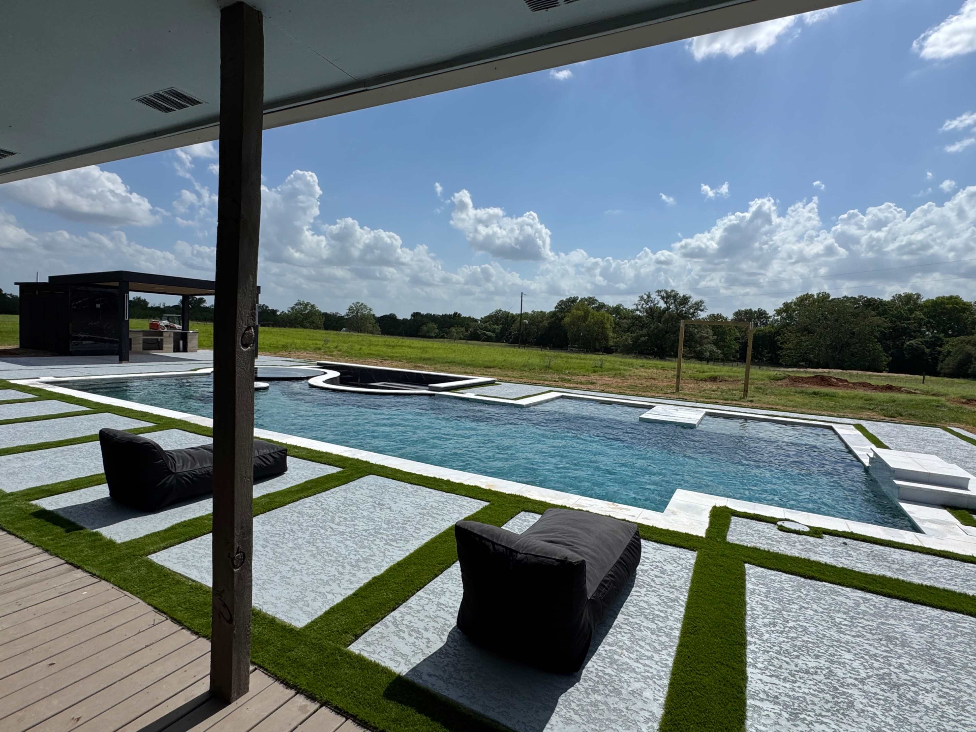 The image shows a modern swimming pool surrounded by landscaped grass and black bean bag chairs, with a lush green field and blue sky in the background.