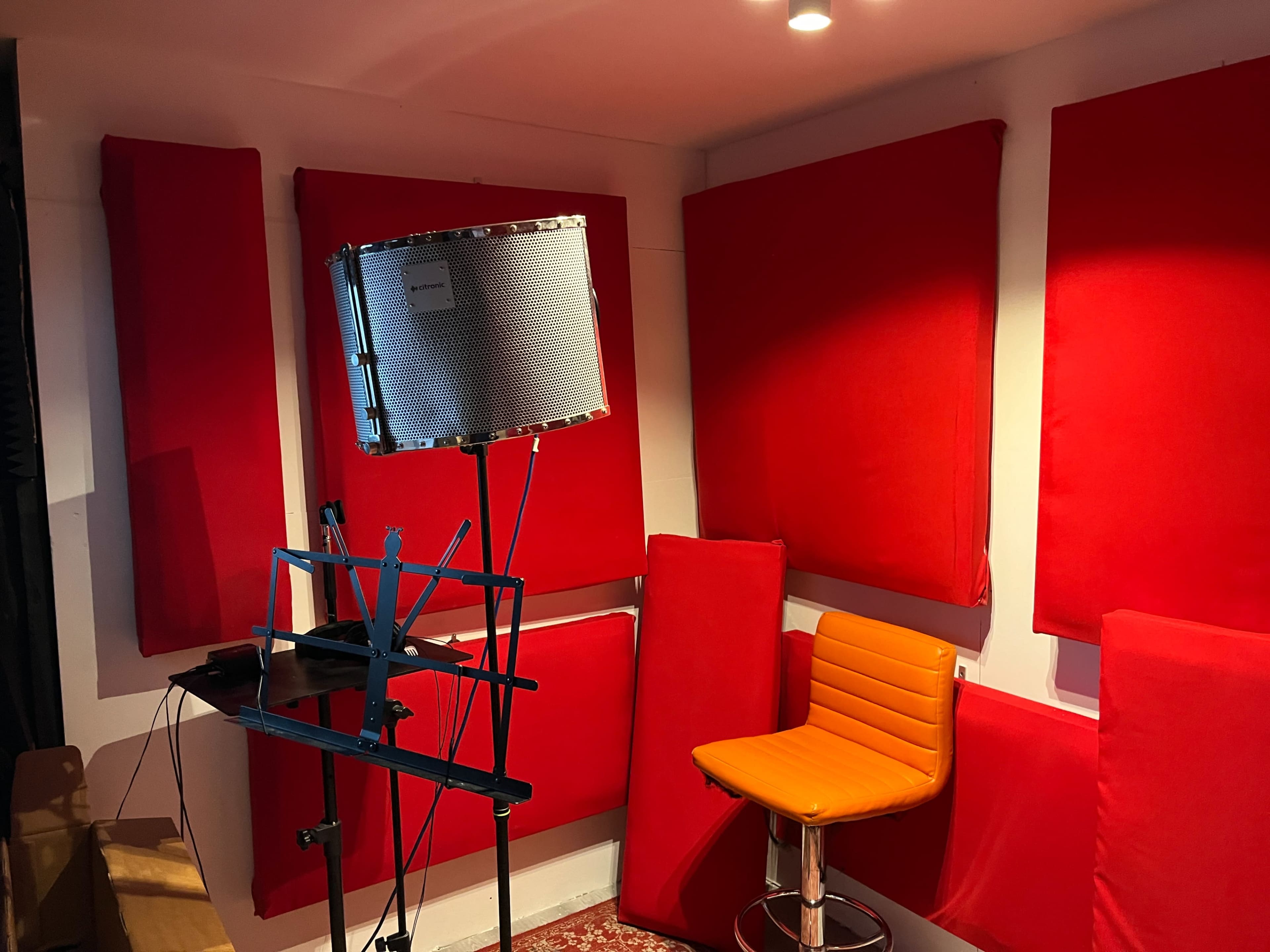 A small recording studio with red soundproofing panels on the walls, a microphone on a stand, a music stand, and an orange chair.