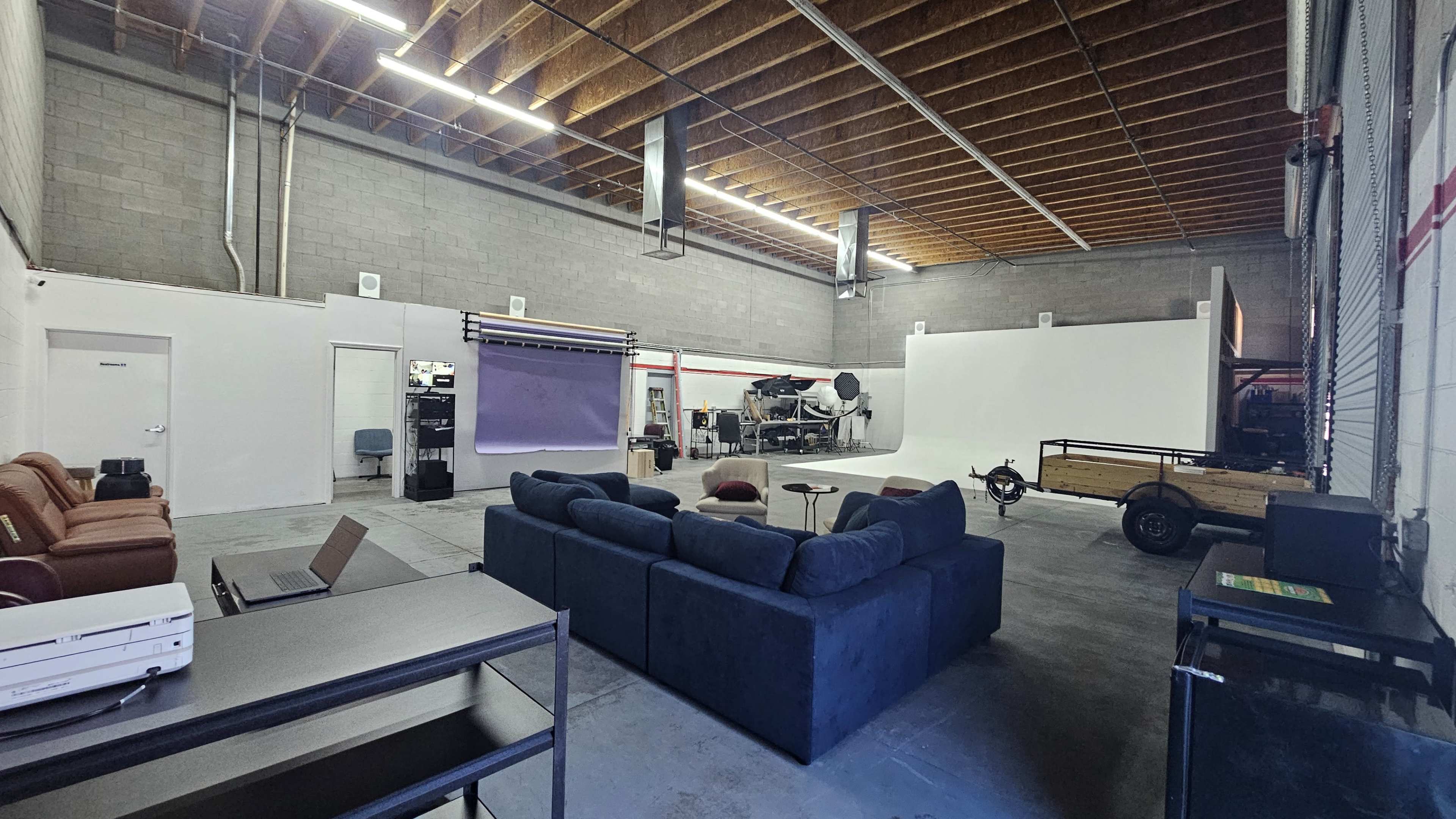 The image shows a spacious studio with a blue couch, a table, a few workstations, and a large backdrop area with various equipment set up.