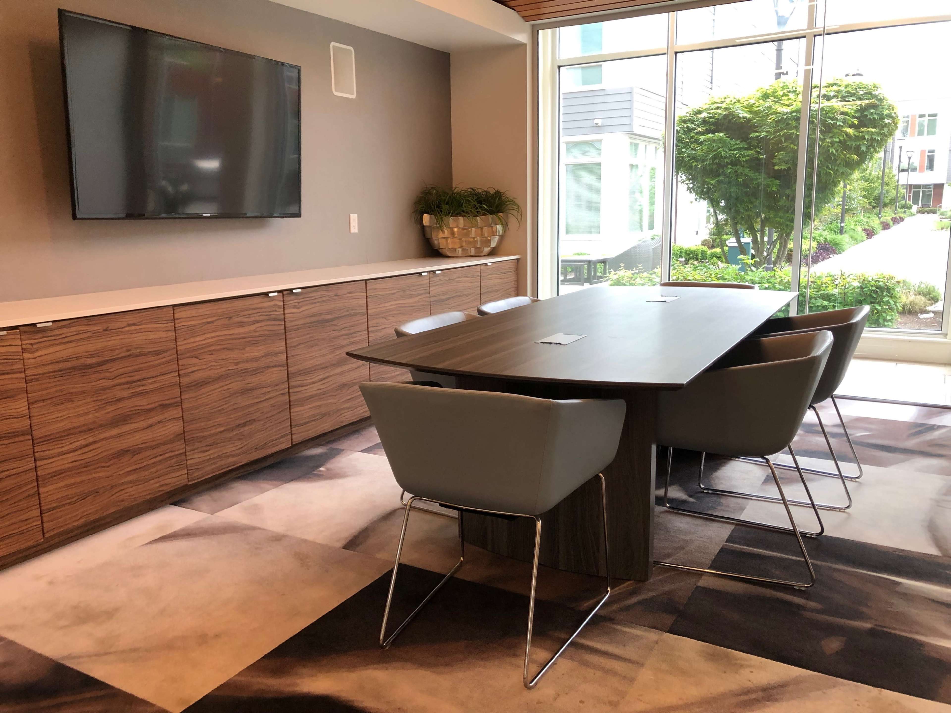 A modern conference room features a large wooden table surrounded by gray chairs, with a wall-mounted TV and large windows overlooking greenery.