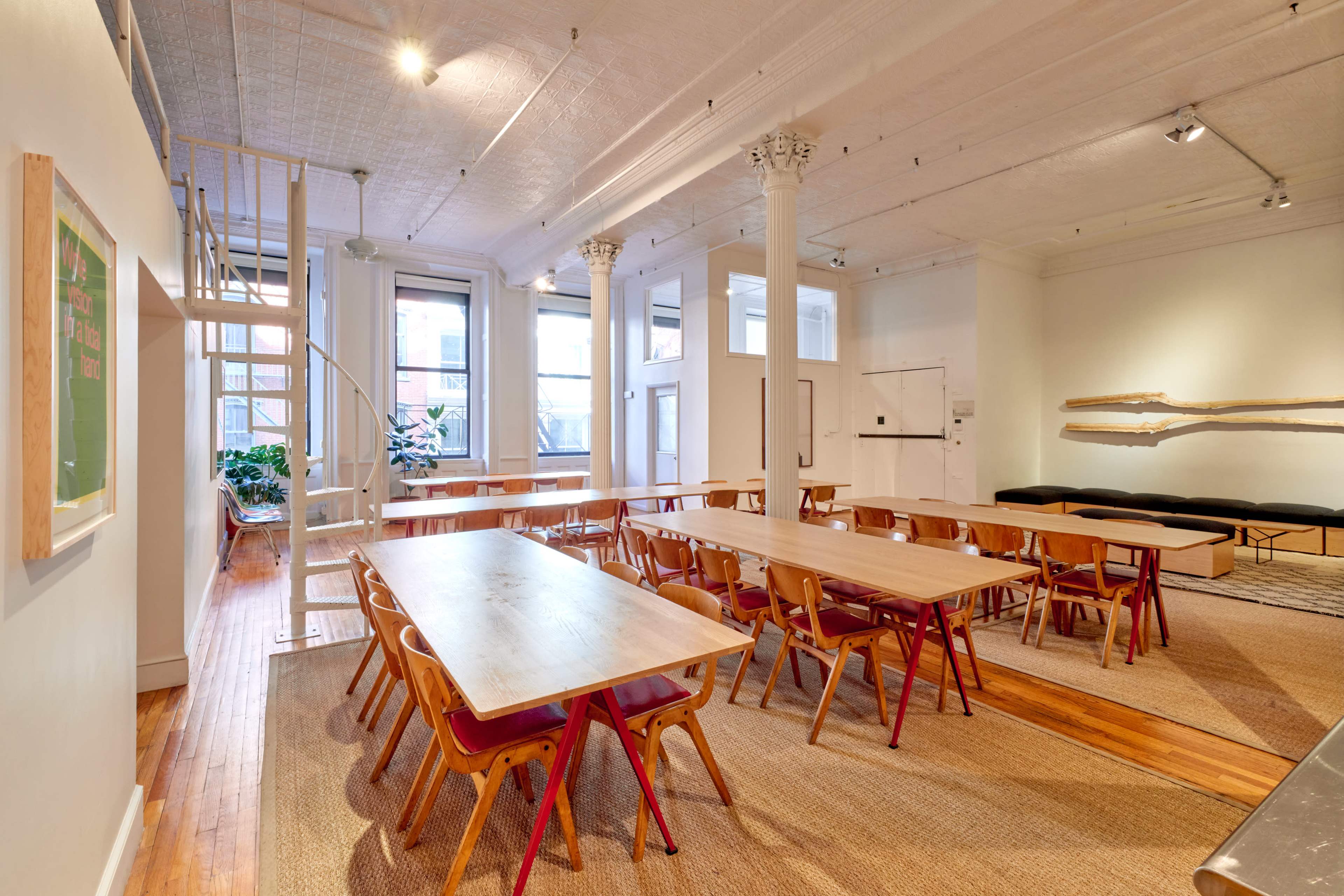 The image shows a spacious, airy room with multiple wooden tables and chairs arranged, large windows allowing natural light in, and a staircase leading to a lofted area.