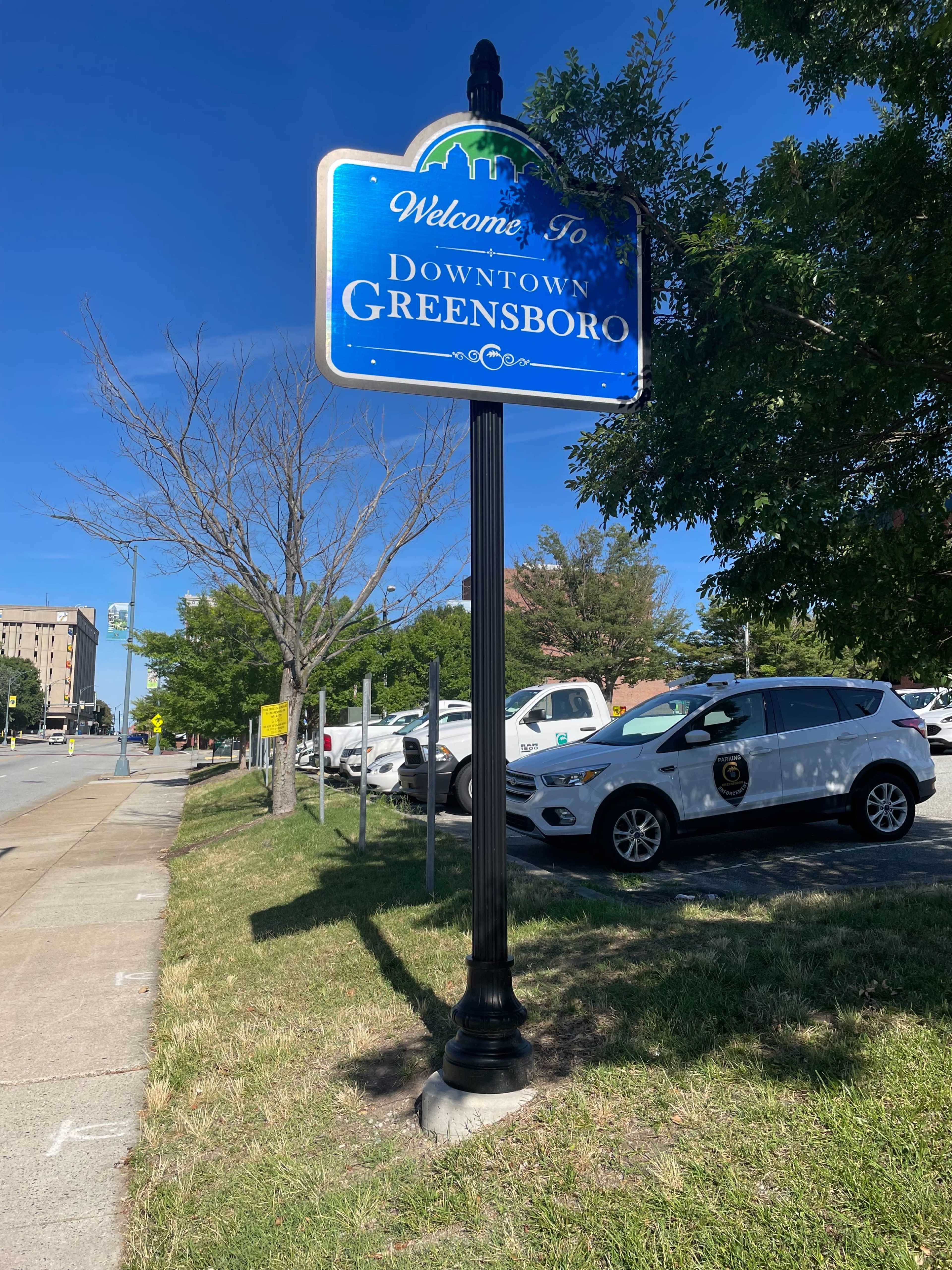 A welcome sign for Downtown Greensboro stands on the side of a road, with parked vehicles in the background.