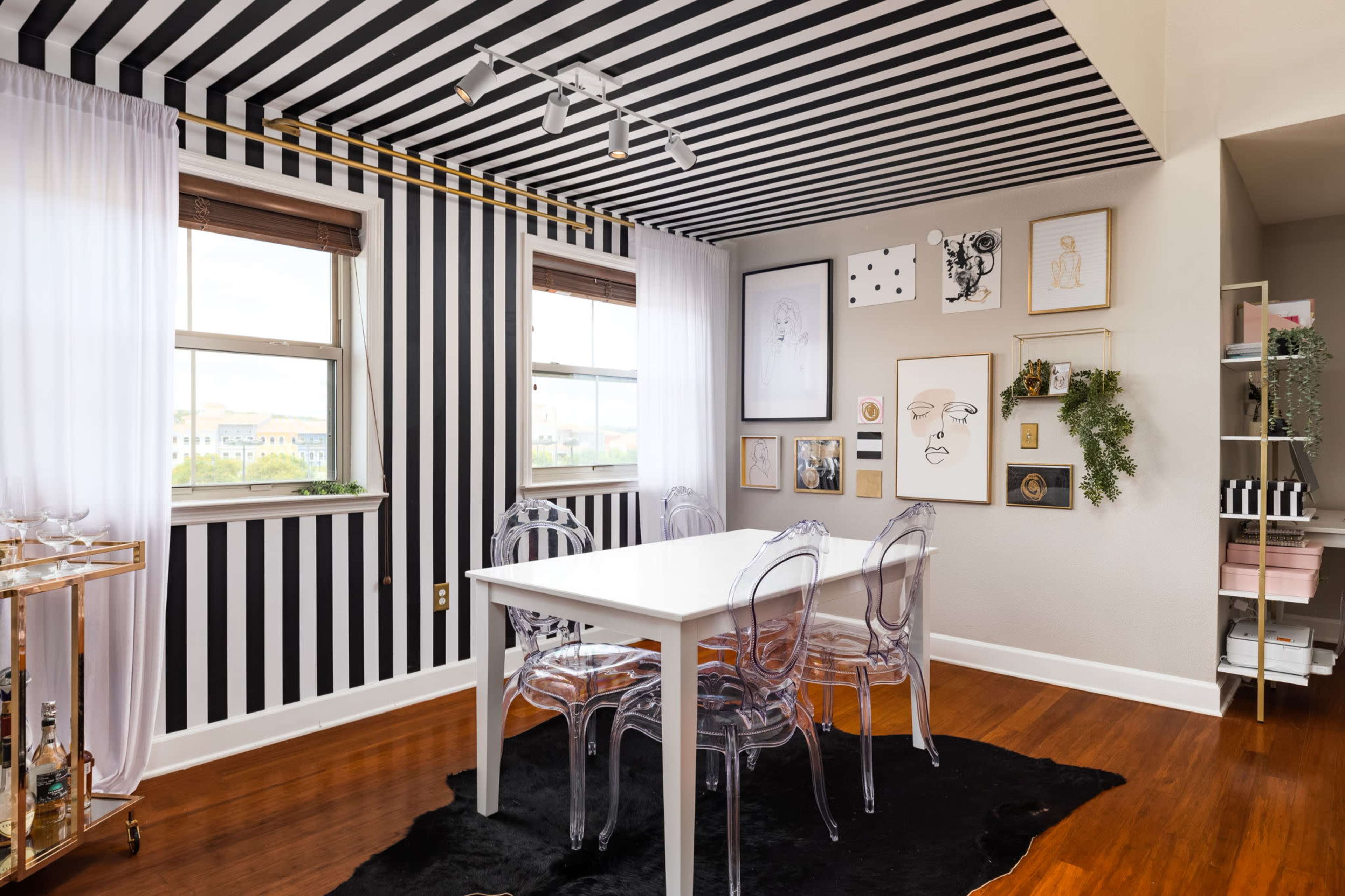 The image shows a dining area with a white table surrounded by clear chairs, set against a bold black-and-white striped wall and ceiling, adorned with various framed artworks.