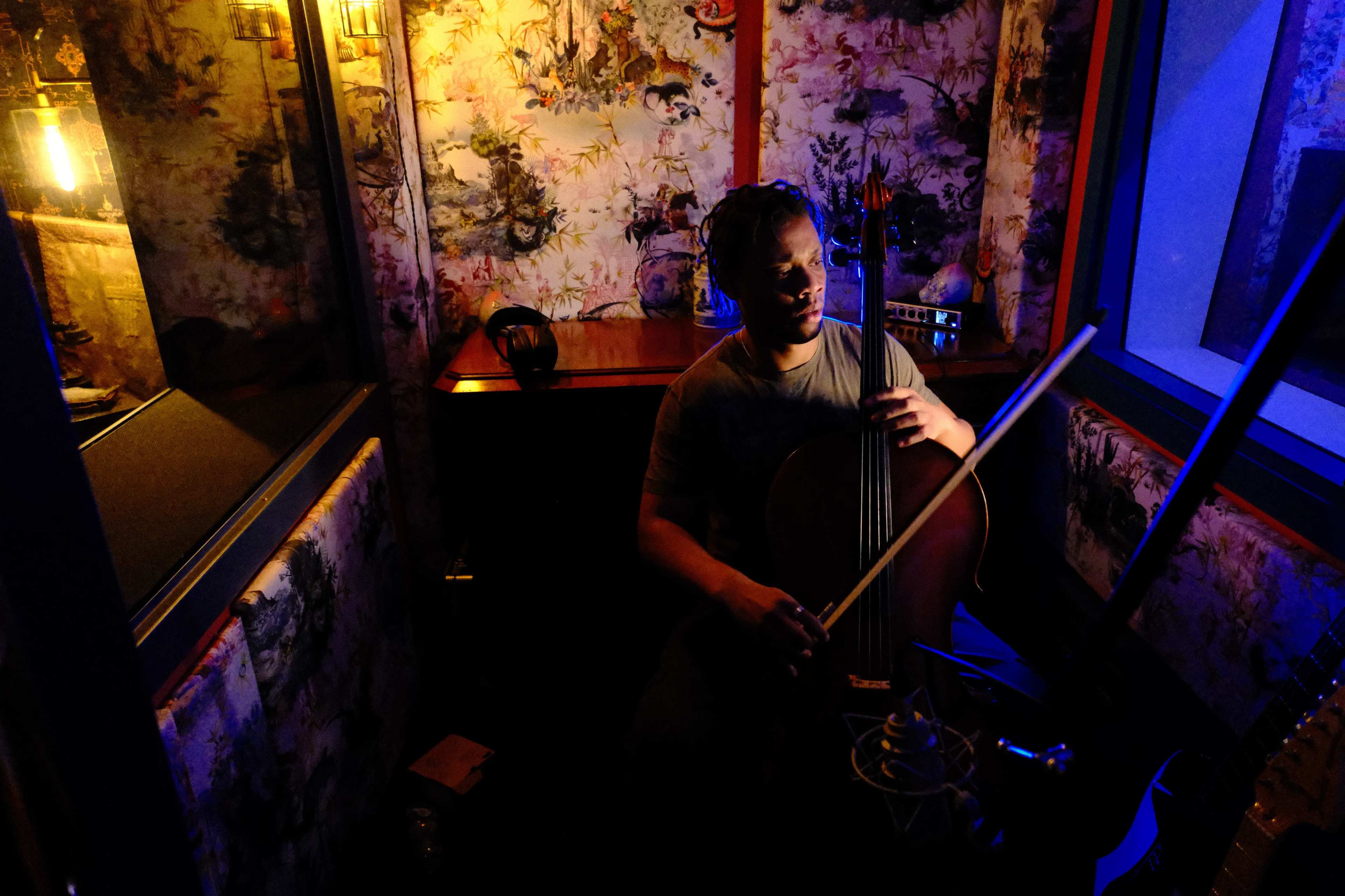 A musician plays a cello in a dimly lit recording studio surrounded by colorful wallpaper.