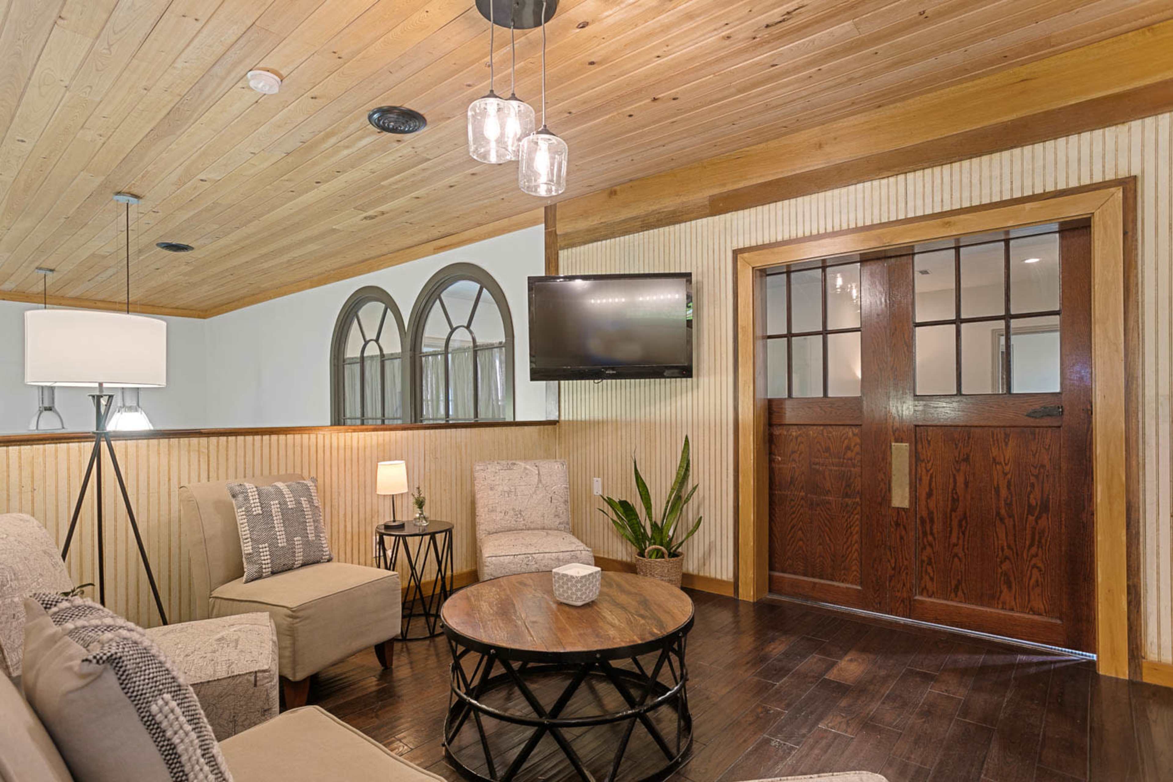 The image shows a living area with two armchairs, a circular coffee table, a flat-screen TV mounted on the wall, and a wooden front door with windows.