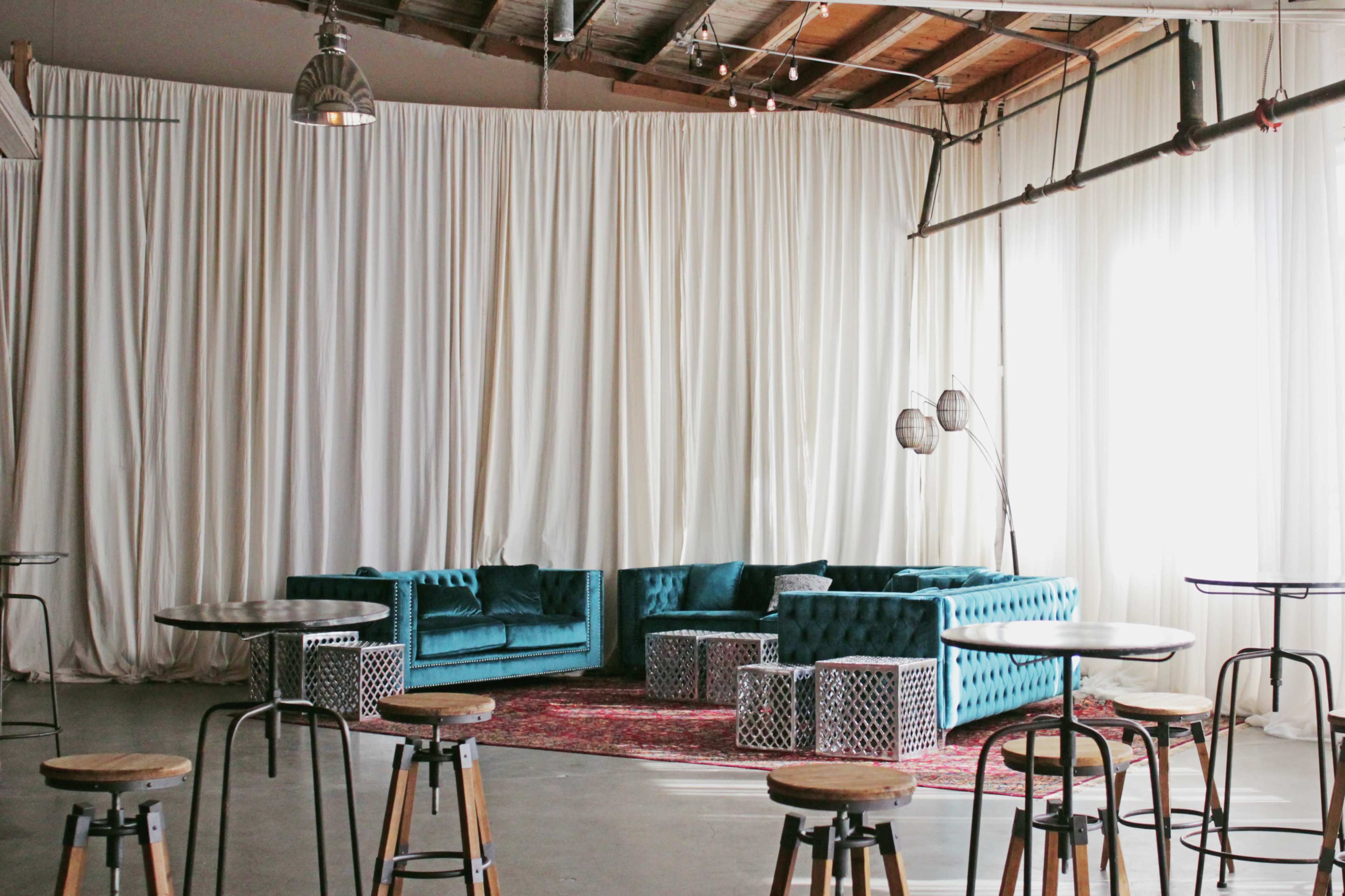A spacious indoor area features turquoise couches, a patterned rug, and high tables with wooden stools against a backdrop of white drapes.