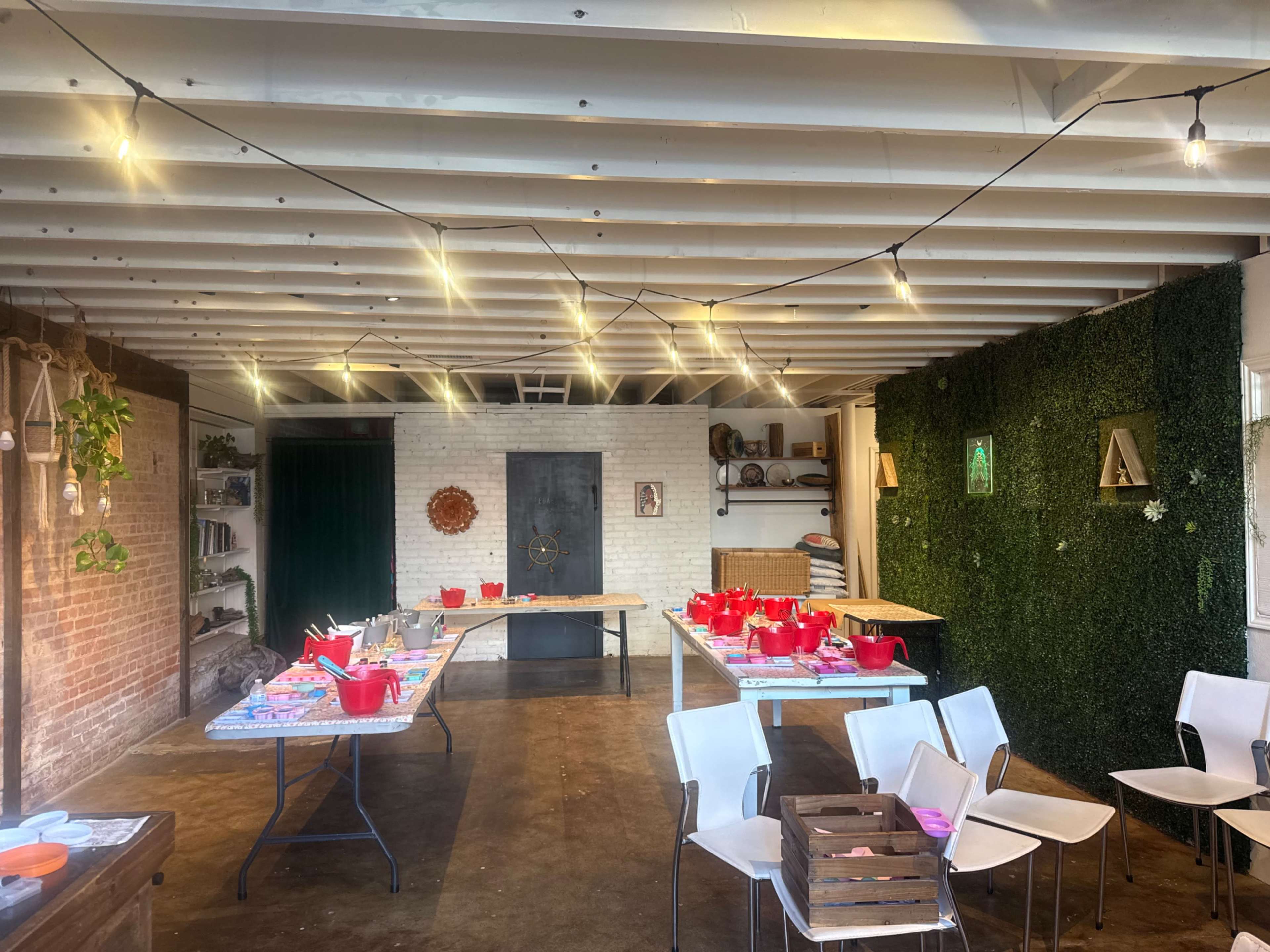 The image shows a neatly arranged indoor space with tables covered in red containers, surrounded by plants, a green wall, and overhead string lights.