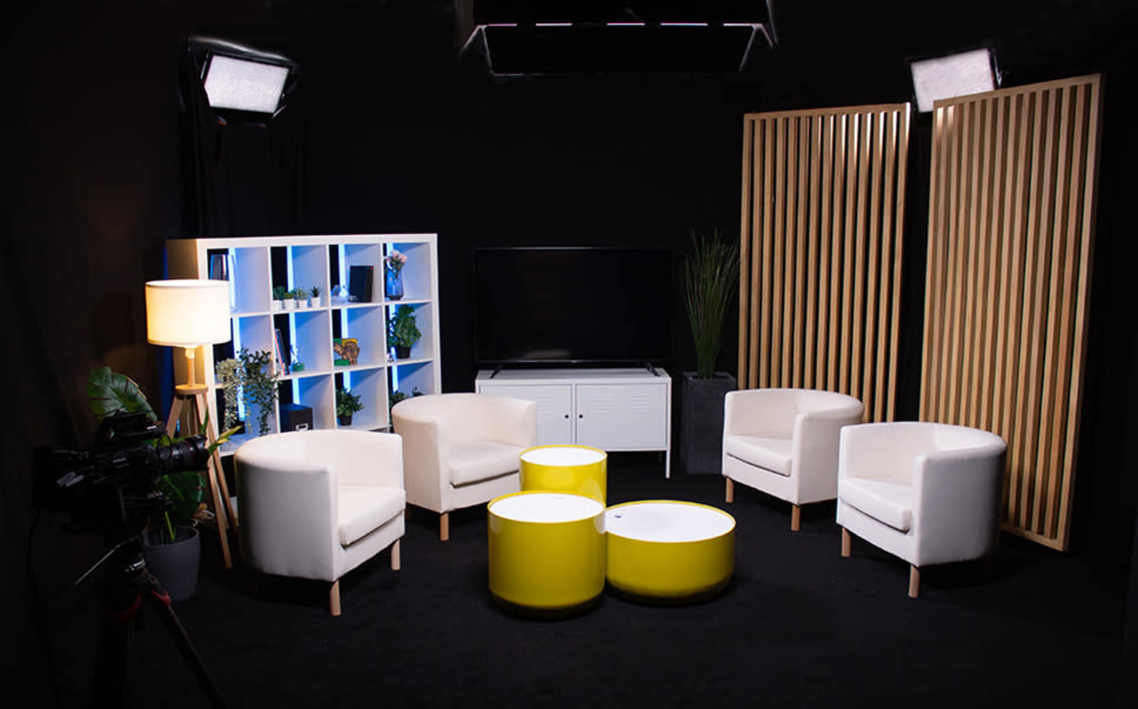 A modern studio setting with four white chairs arranged around two yellow coffee tables, a bookshelf, and a television on a low cabinet, all under soft lighting against a black backdrop.