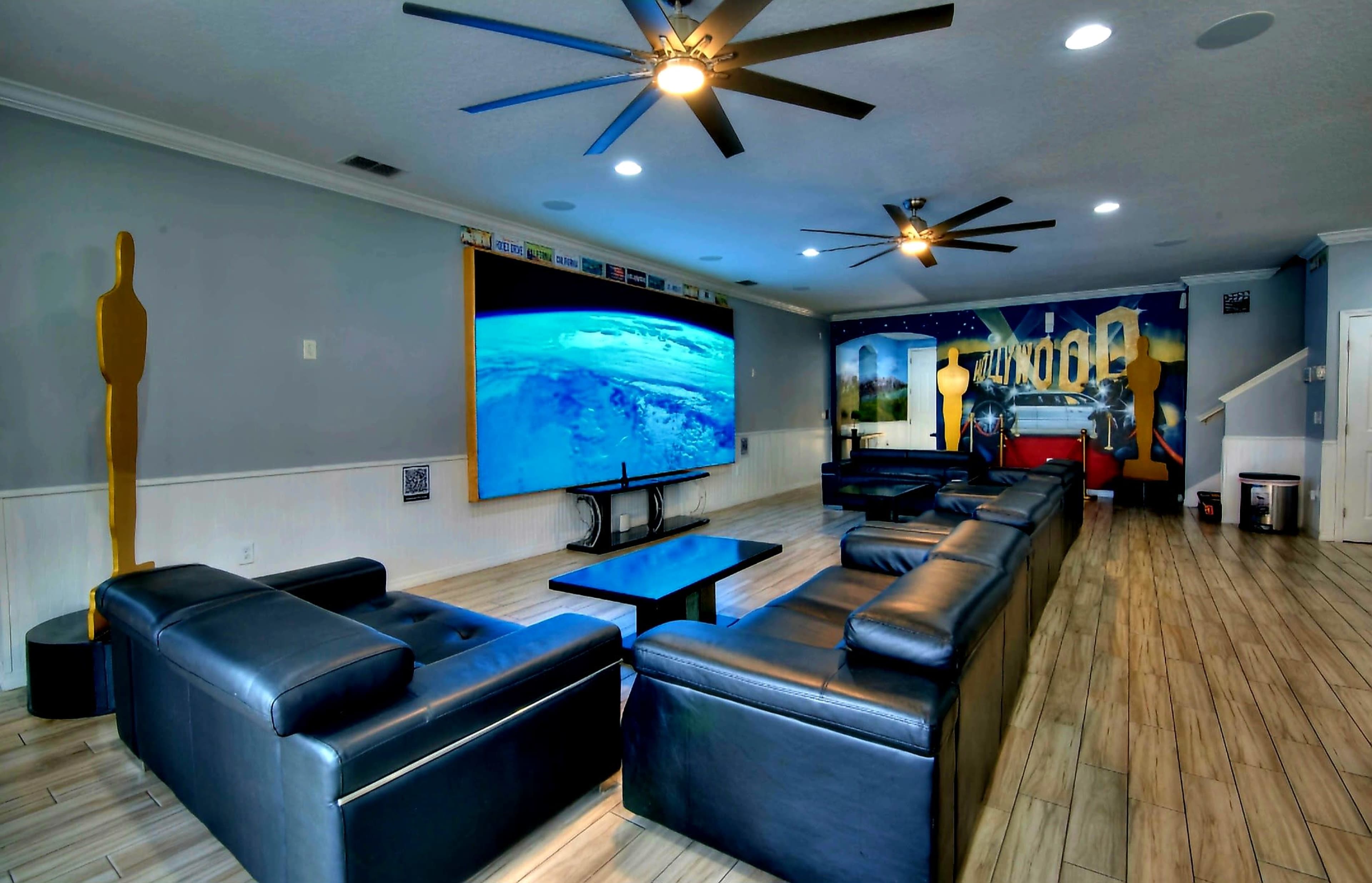 A large living room features black leather sofas, a central coffee table, and a wall-mounted screen displaying a scenic view, with decorative elements in a modern design.