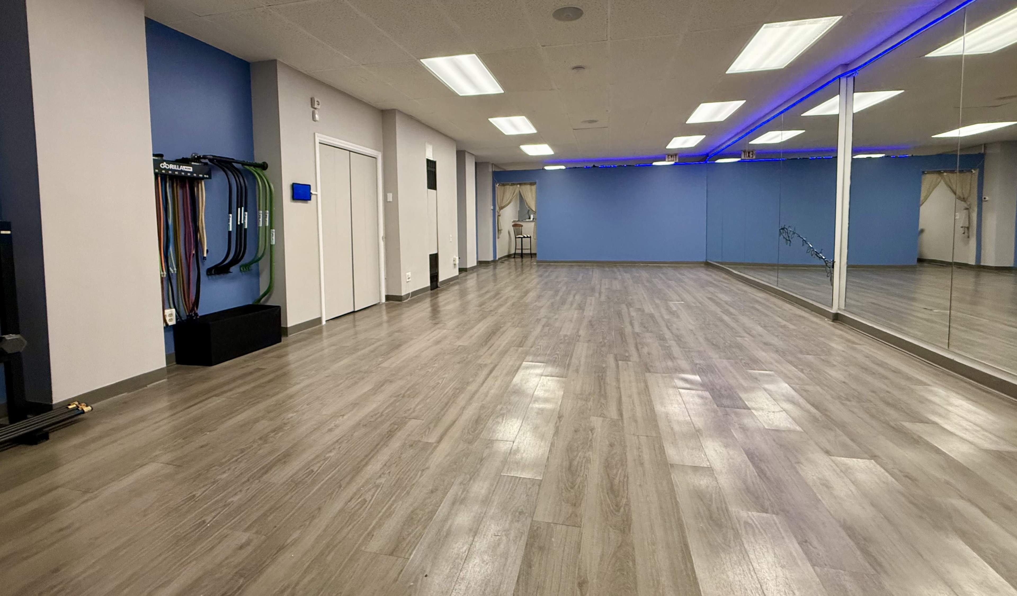 The image shows a spacious, well-lit exercise studio featuring mirrored walls and wooden flooring, with fitness equipment hung on the wall.