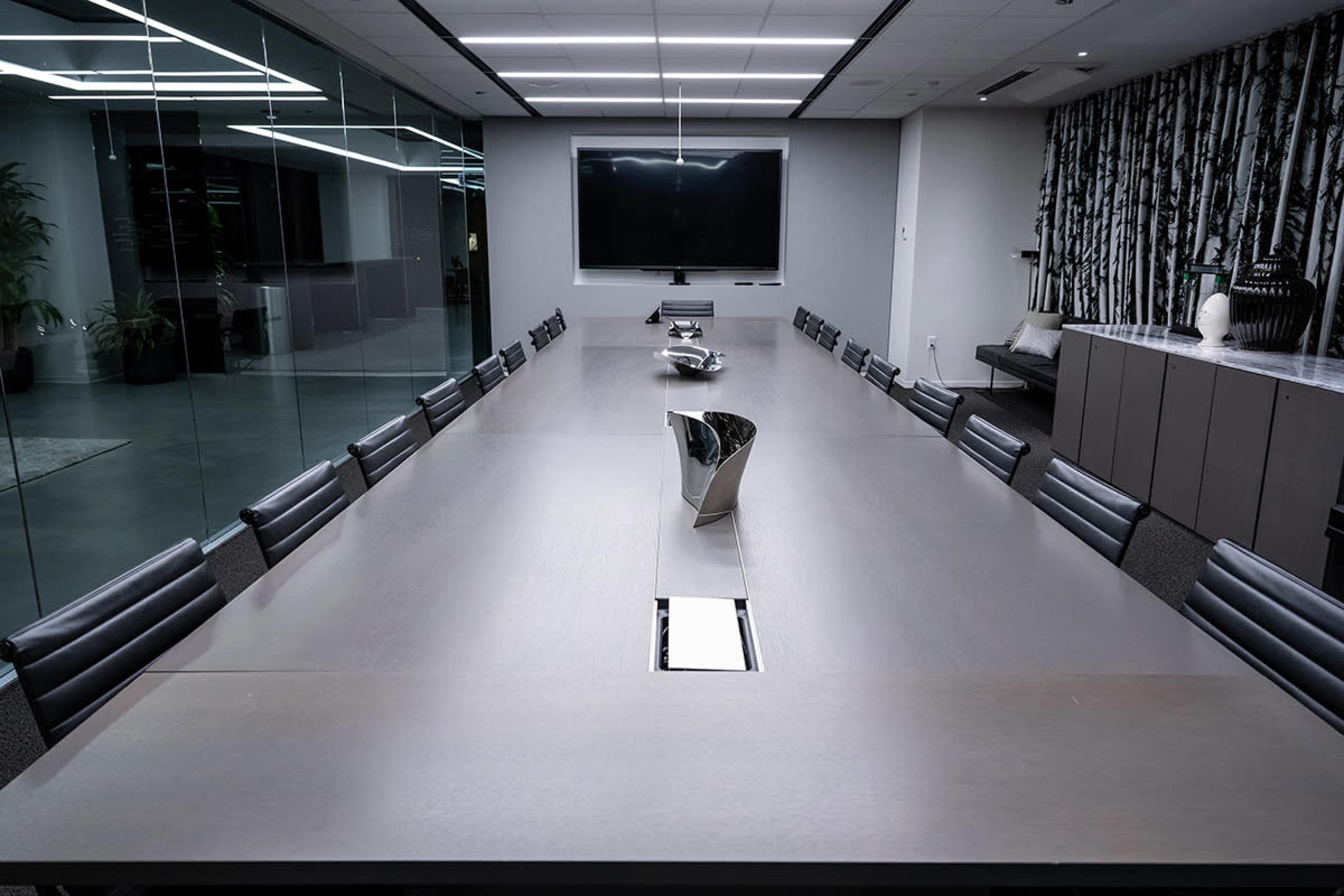 A large, modern conference room features a lengthy table surrounded by black leather chairs, with a television mounted on the wall and a decorative screen in the background.