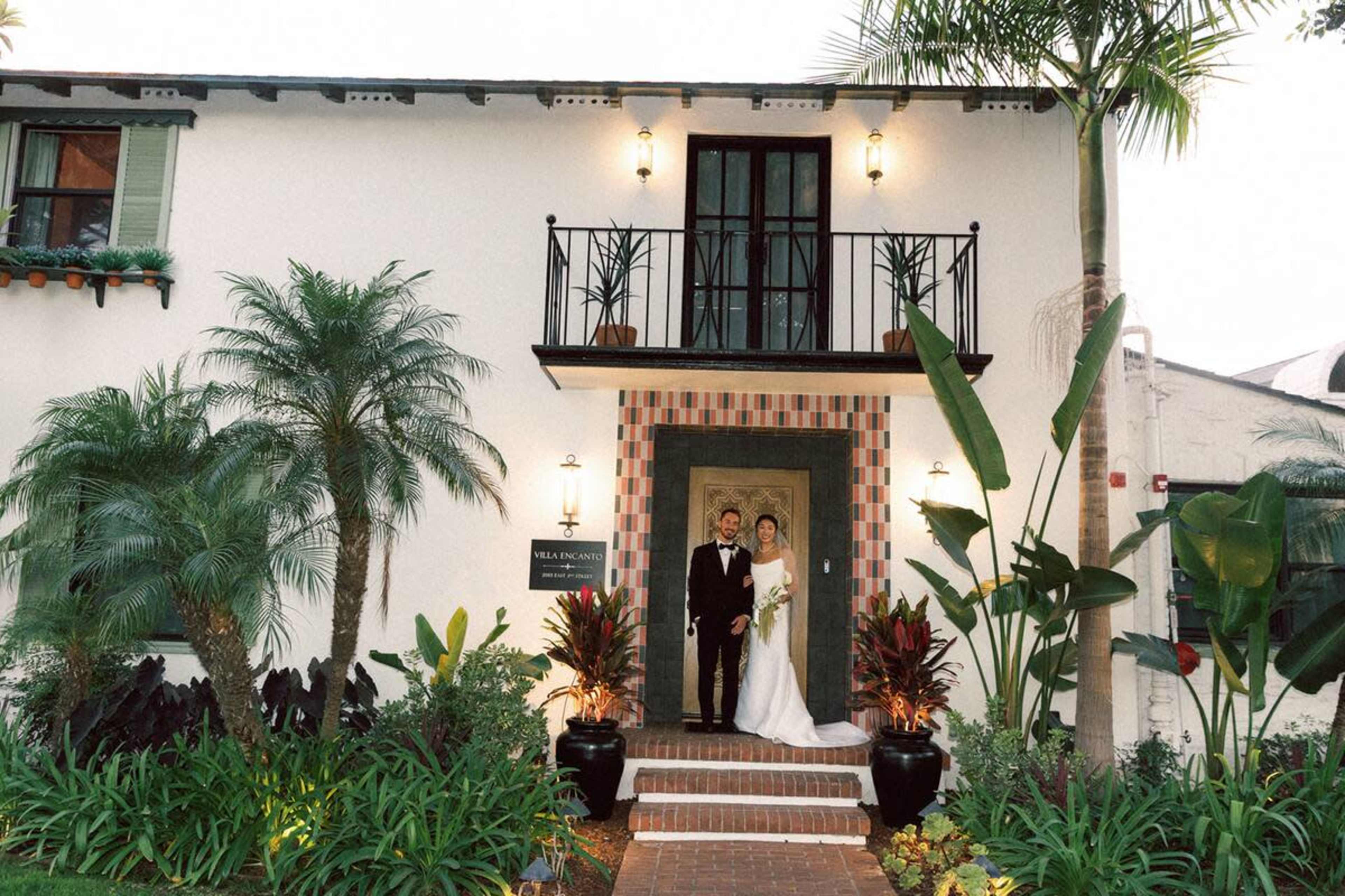 A newlywed couple stands together on the steps of a decorative entrance to a building surrounded by tropical plants.
