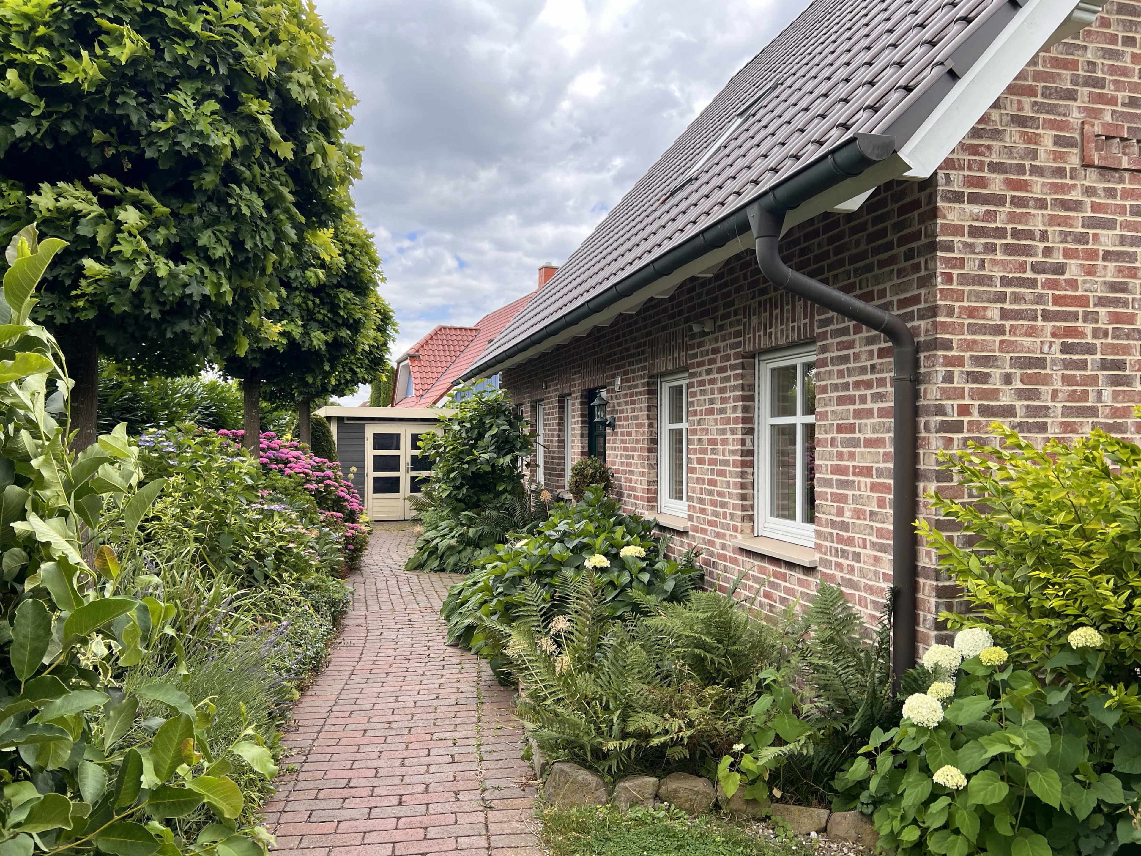 A brick house is flanked by a walkway lined with vibrant plants and bushes leading to a nearby structure.