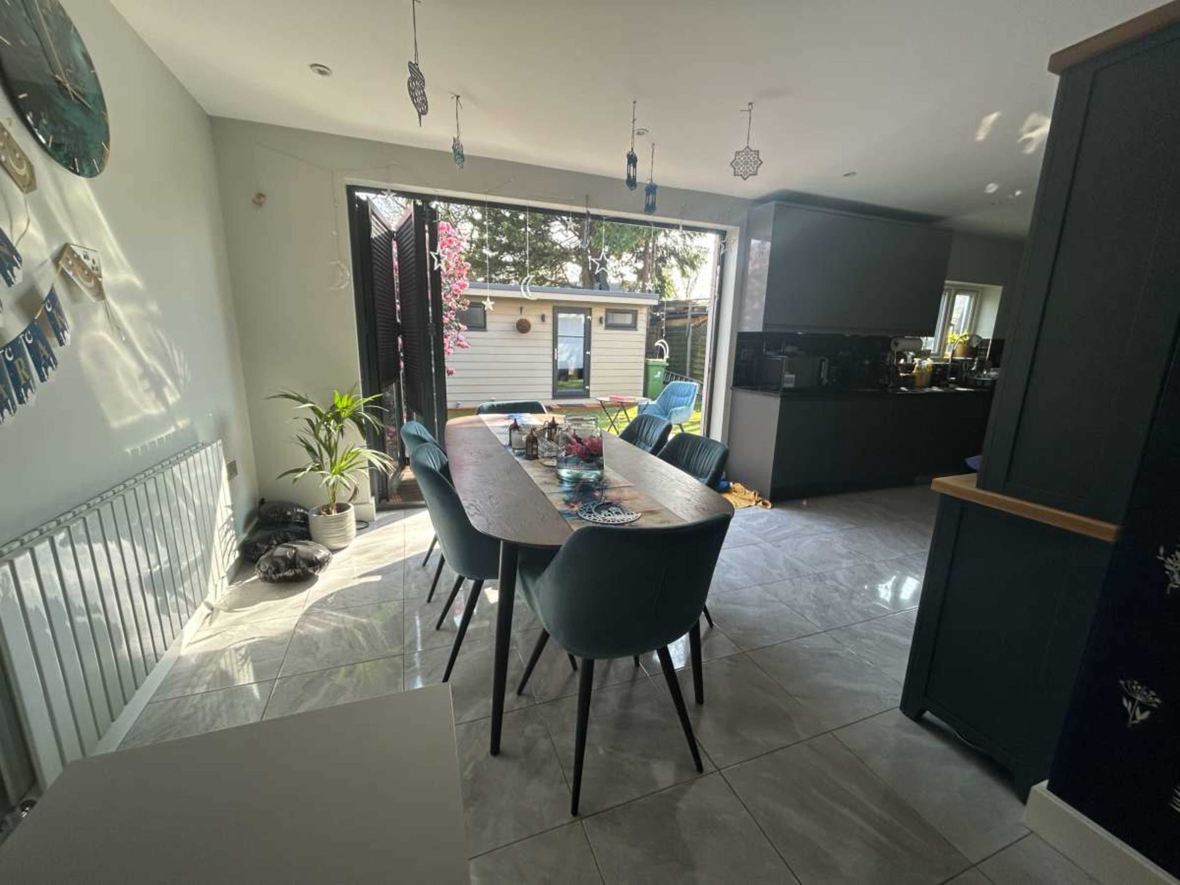 A modern dining area features a long table surrounded by blue chairs, with sunlight streaming in through open sliding doors that lead to a patio.