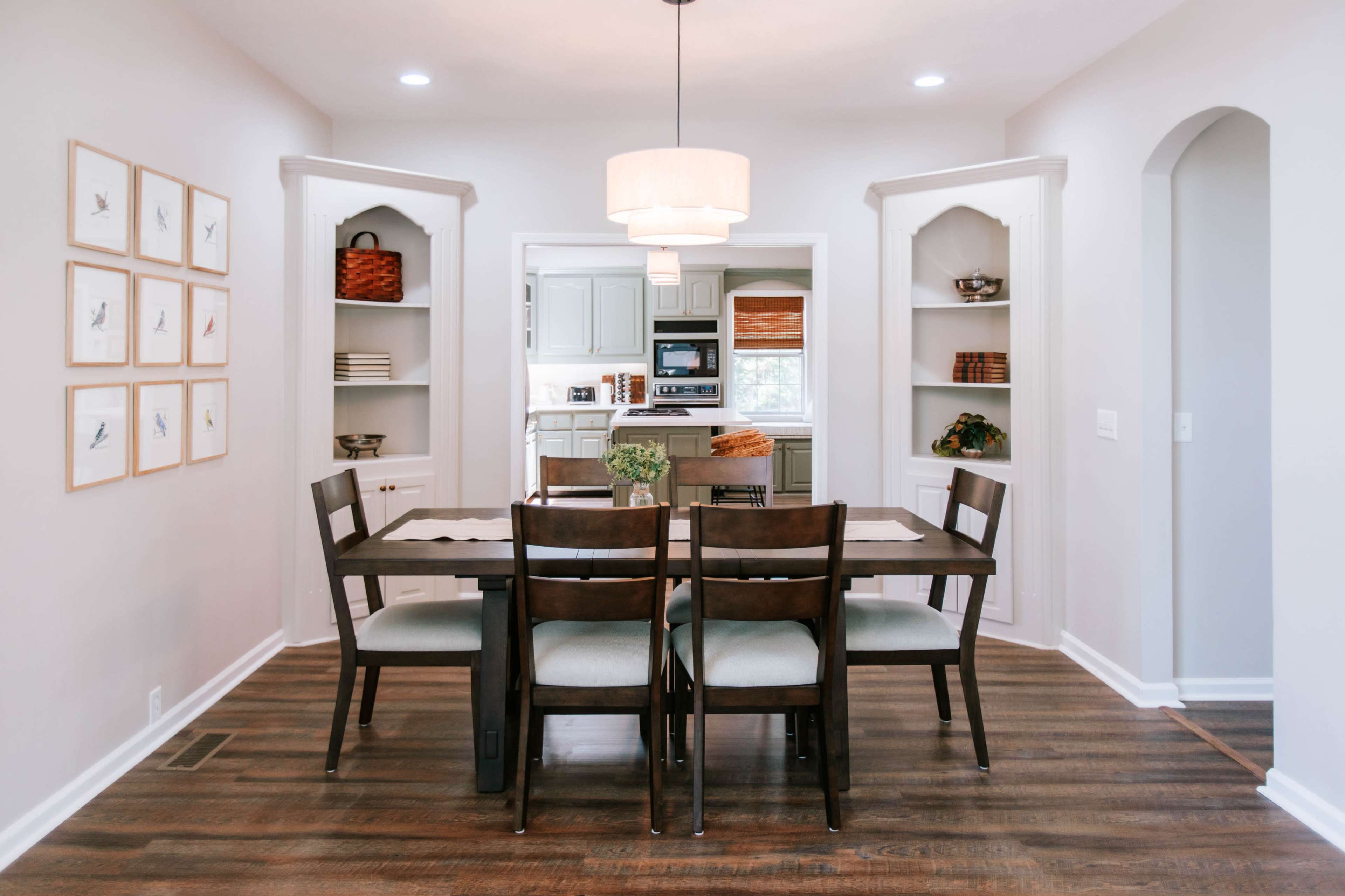The image shows a well-lit dining area with a wooden table and chairs, framed bird prints on the wall, and open shelves displaying decorative items.
