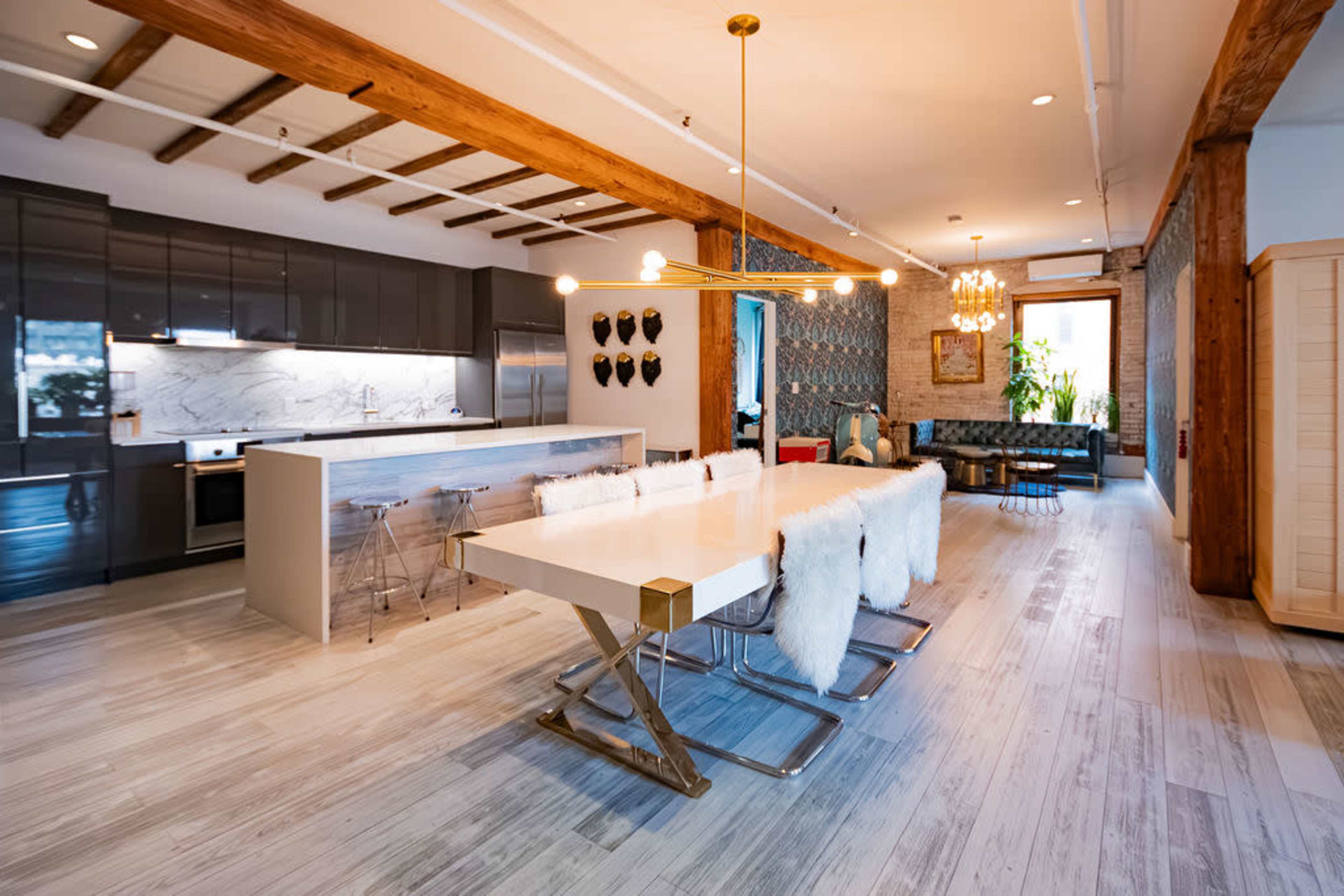The image shows a modern dining area with a long white table and plush chairs, adjacent to a contemporary kitchen featuring dark cabinetry and a marble backsplash.