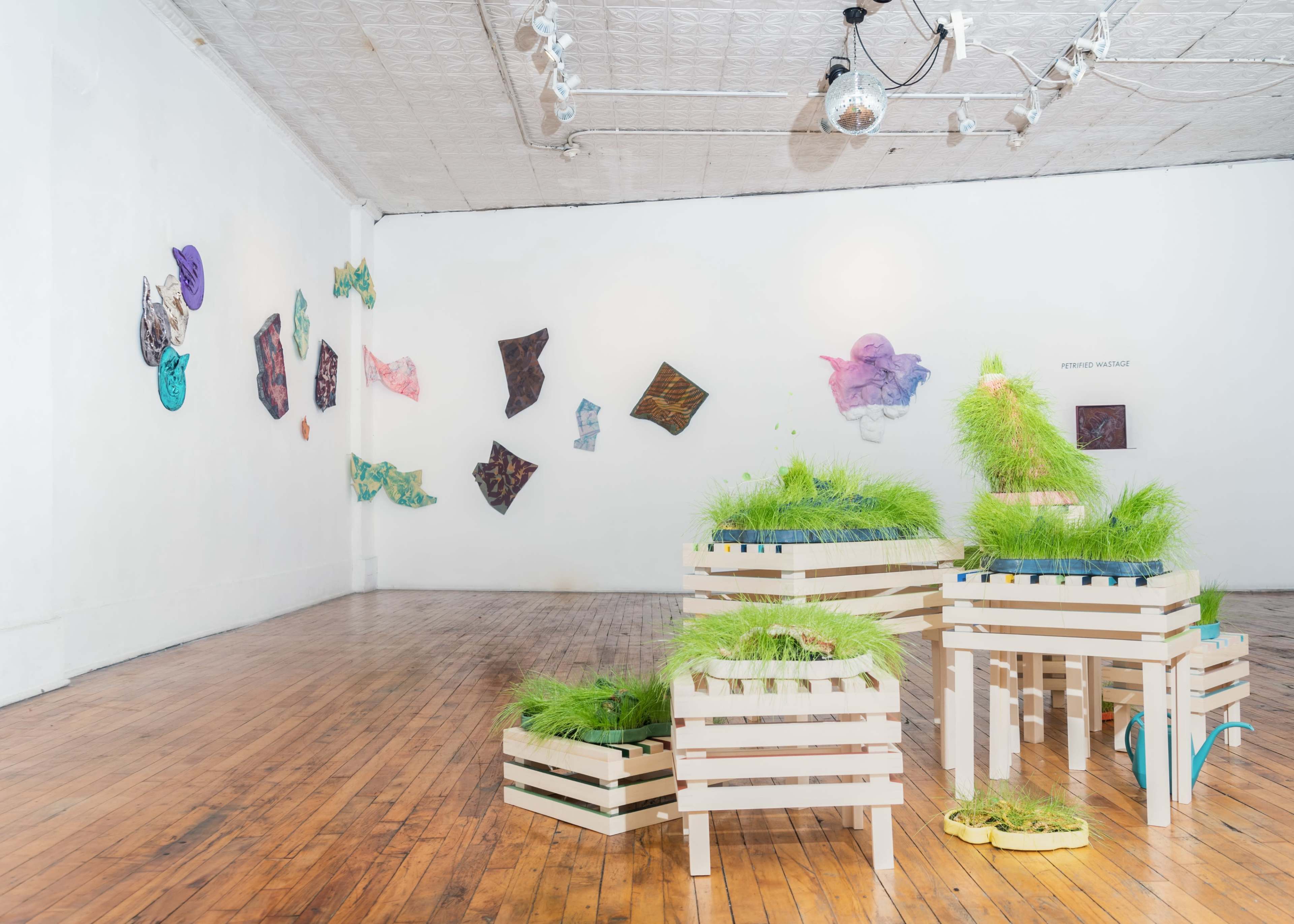 An art gallery with wooden crates filled with grass and colorful fabric artworks displayed on the walls.