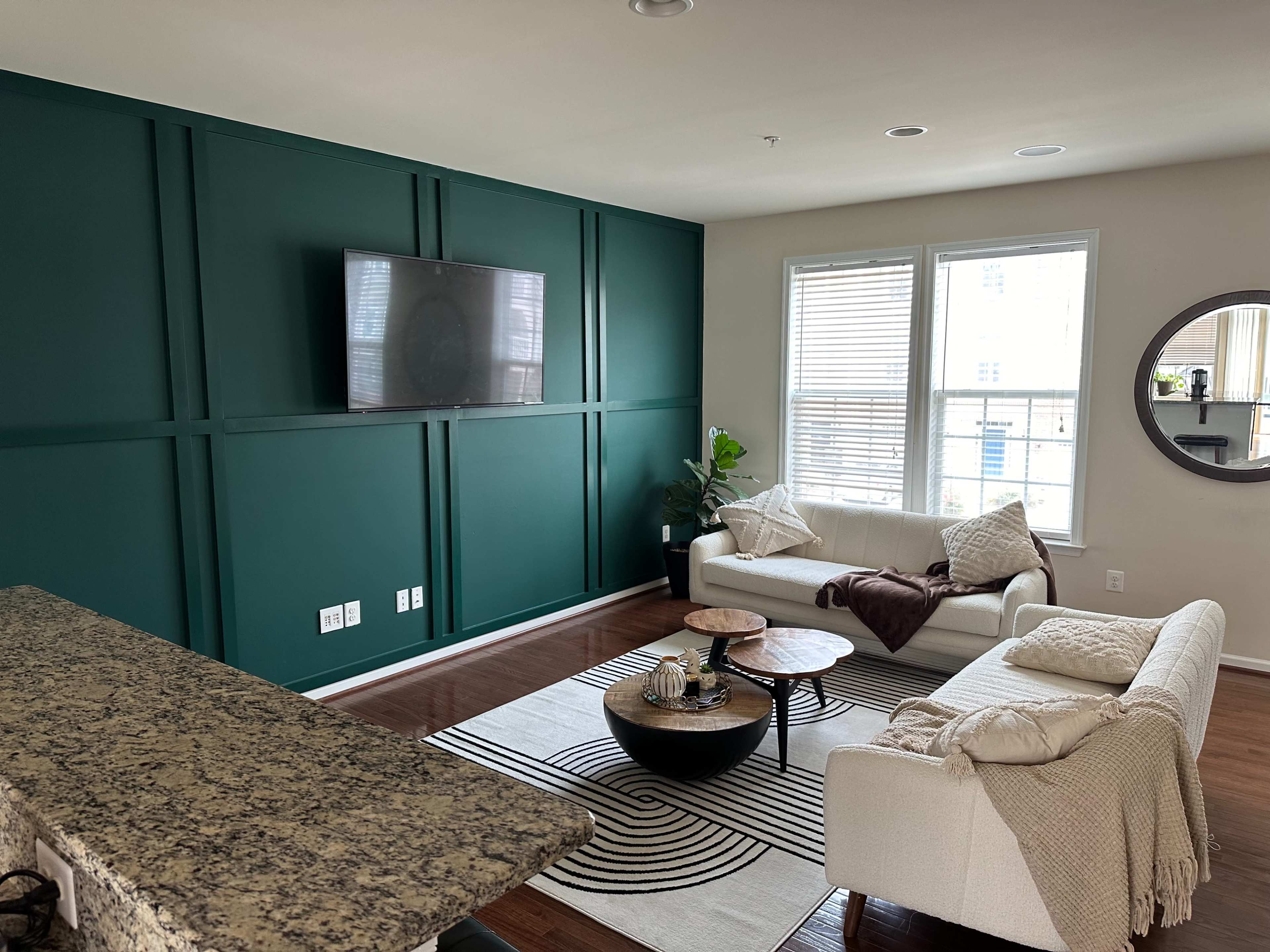 The living room features a green accent wall with a mounted TV, two white sofas, a circular coffee table, and large windows allowing natural light.