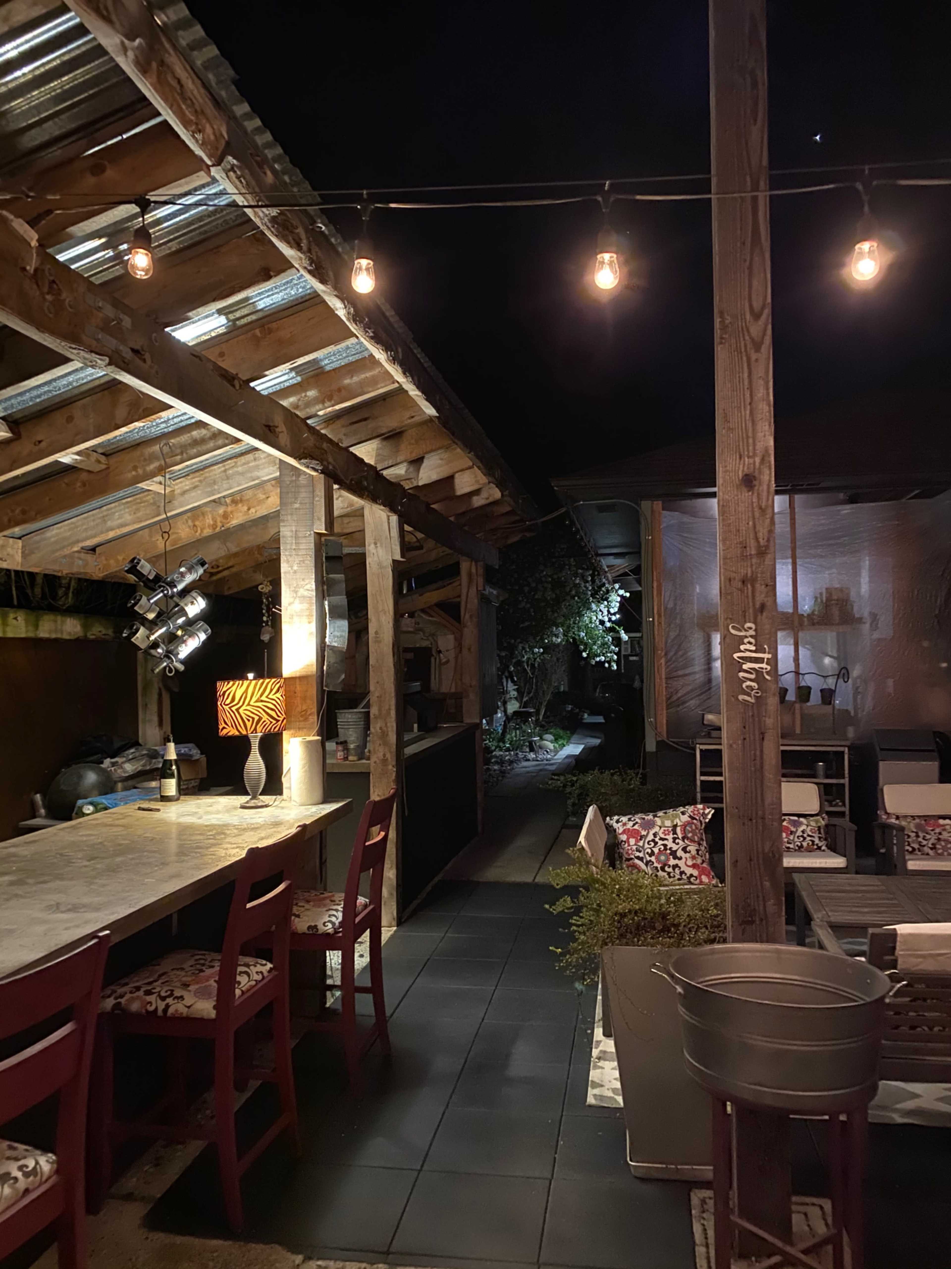 A dimly lit outdoor patio features a bar with high stools, string lights overhead, and a pathway leading to a garden area.