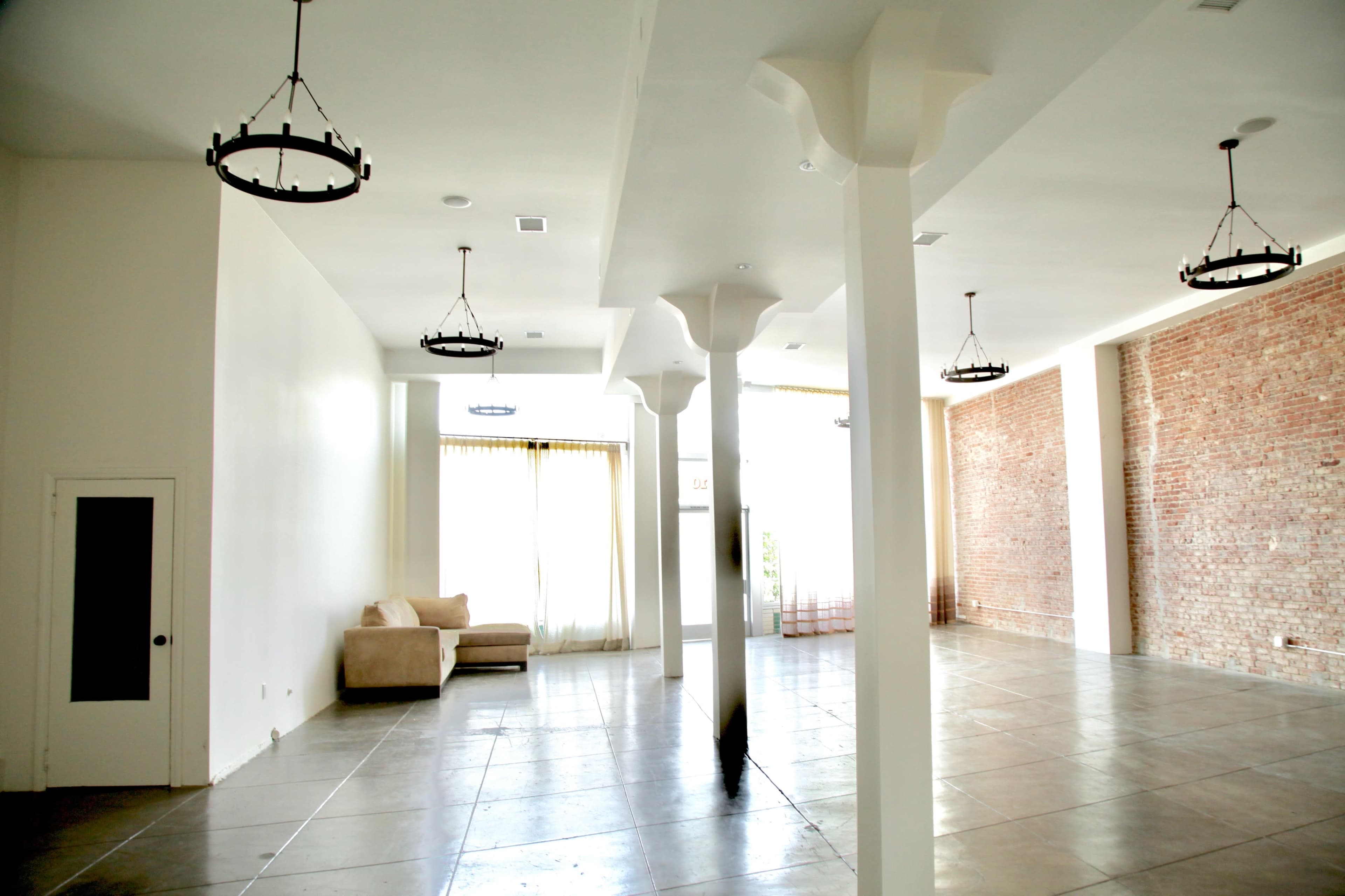 The image shows a spacious, empty room with high ceilings, large windows, and exposed brick walls.
