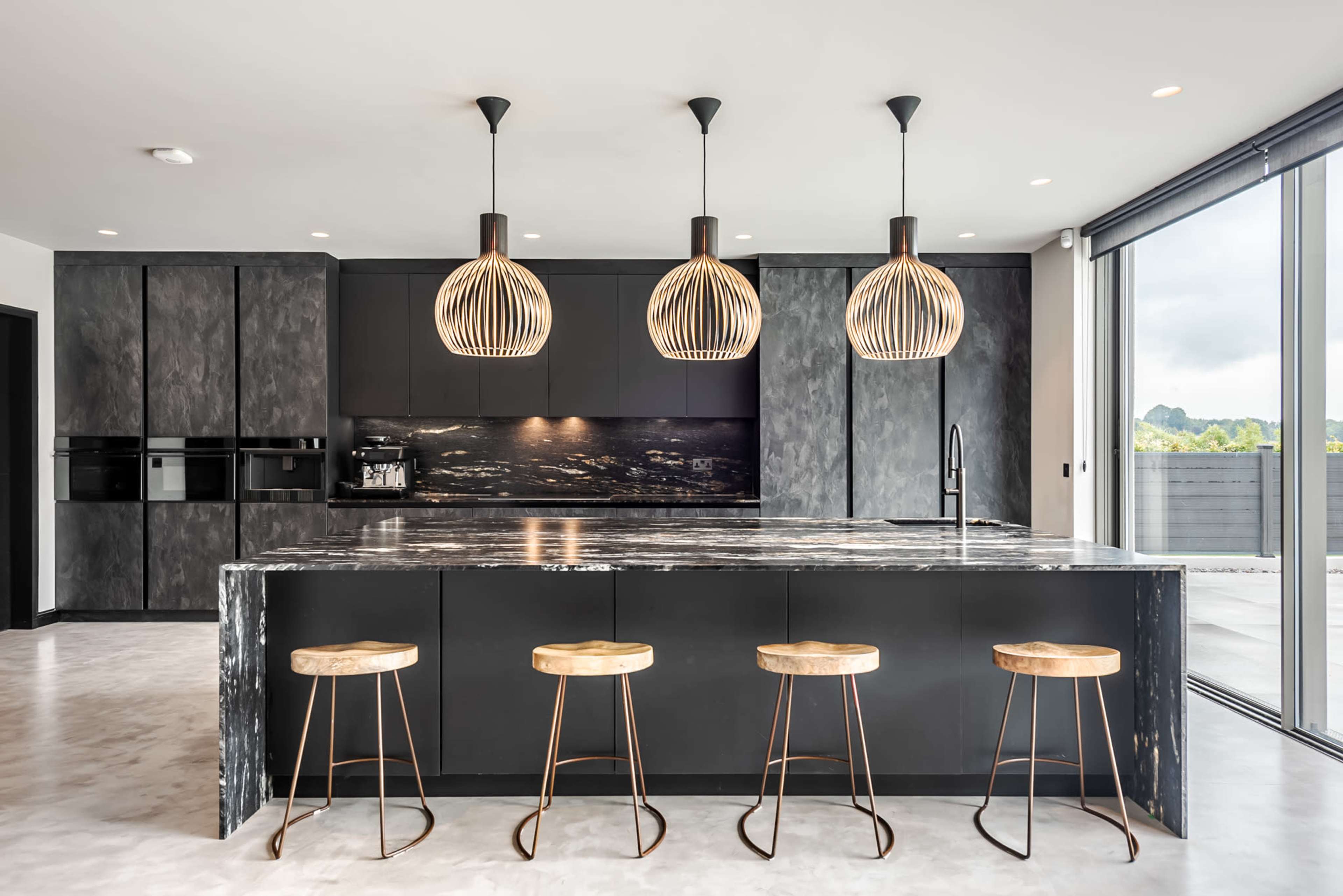 A modern kitchen featuring a dark cabinetry layout, a large island with a marble countertop, and three pendant lights above it, accompanied by four wooden stools.