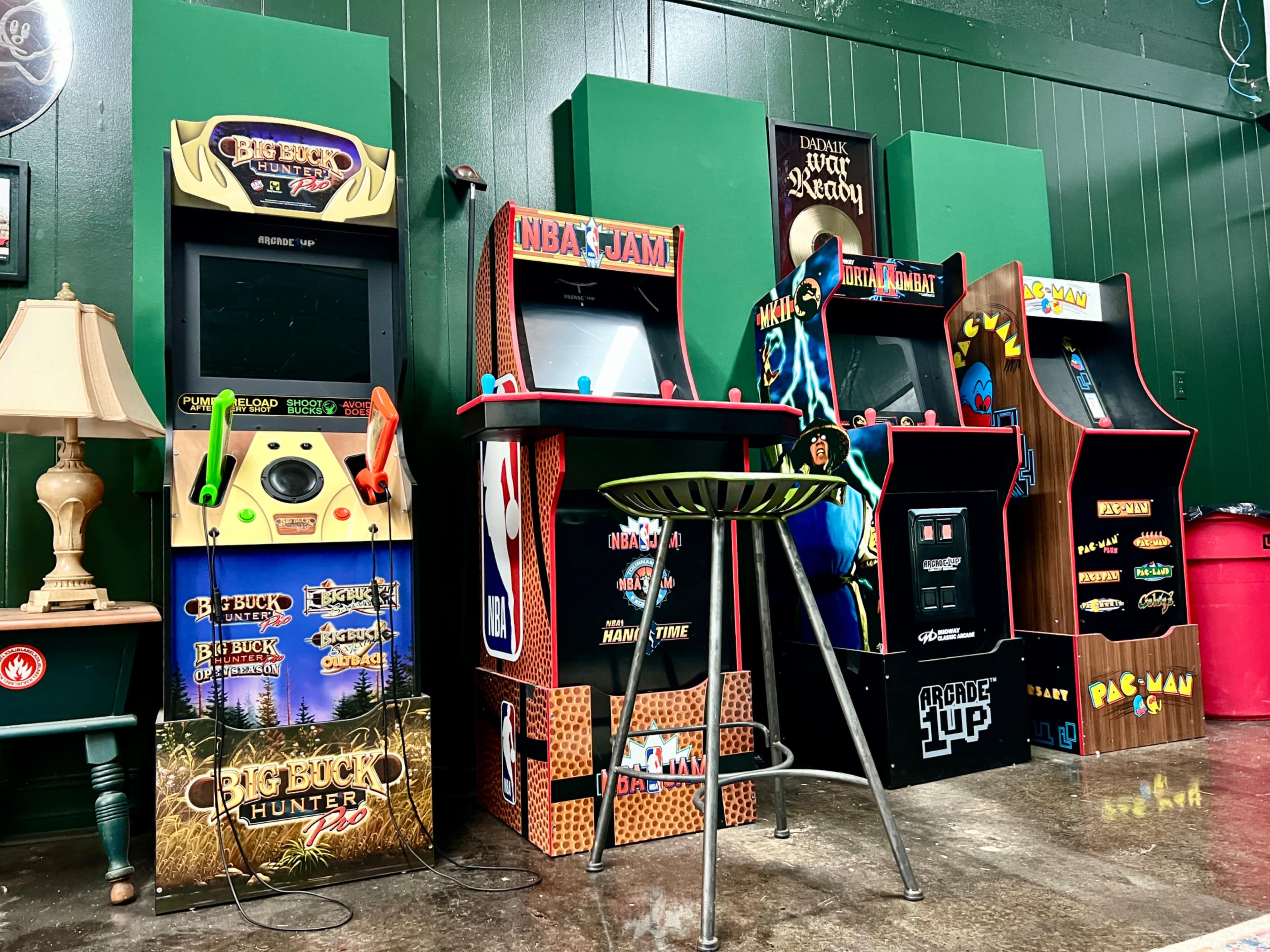 The image shows a row of vintage arcade game machines, including "Big Buck Hunter," "NBA Jam," and "Pac-Man," set against a green wall in a game room.